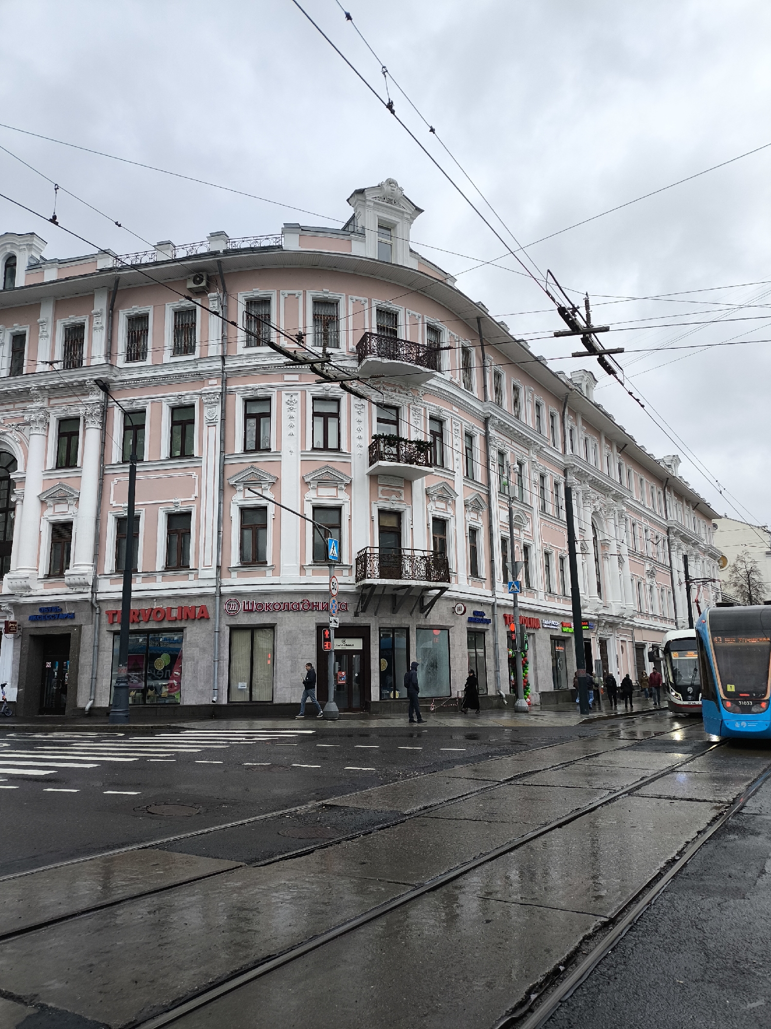 The image depicts a Moscow street scene on an overcast day, characterized by wet asphalt and a grey sky. A prominent, multi-story building with a light pink facade and white ornate architectural details, including columns, window frames, and decorative balconies with dark wrought-iron railings, occupies the left and center. The ground floor features several commercial establishments, notably a "TERVOLINA" shoe store and a "Шоколадница" (Shokoladnitsa) coffee house, both displaying red signage.
A complex network of overhead power lines and tram wires crisscrosses the sky above the building. In the foreground, wet tram tracks run diagonally across the street. A blue and white tram, partially visible on the right, displays the number "31033" on its front. Several pedestrians are seen walking on the sidewalks and crossing the street, dressed in warm jackets and coats, suggesting cool weather. A circular speed limit sign indicating "50" is visible on a pole near the tram tracks. The scene captures typical daily urban activity in a city environment during what appears to be a cool, cloudy daytime.