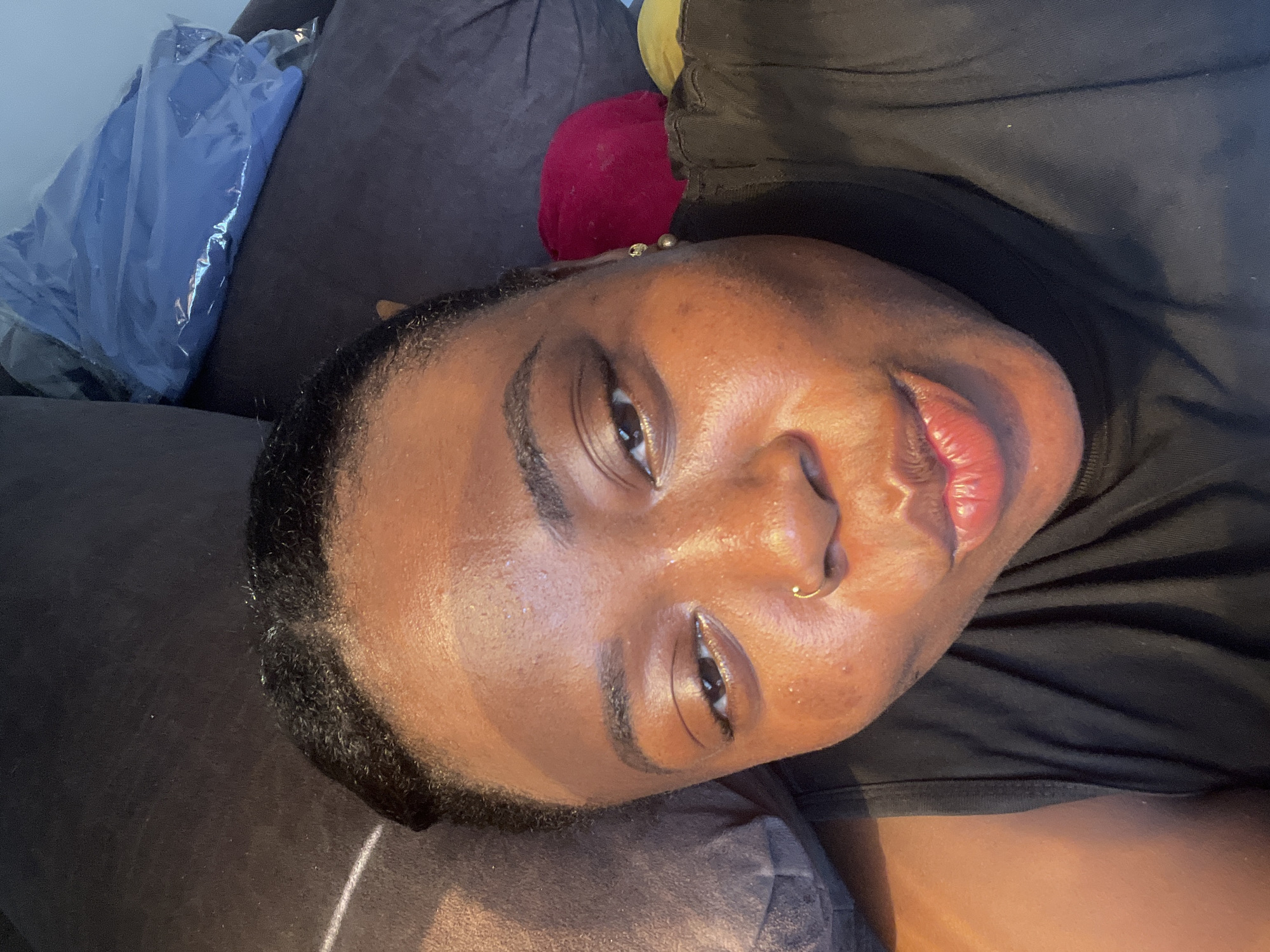 A close-up, downward-facing image shows a person lying on a dark, possibly gray, cushioned surface. The person has dark skin, dark hair, and what appears to be a nose ring. The individual is wearing a black top. Behind the person, elements of the scene include a crinkled blue object, and a reddish rounded item on the cushioned surface. The location is indeterminate, and there is no information to suggest this image was taken in Abuja, Nigeria.