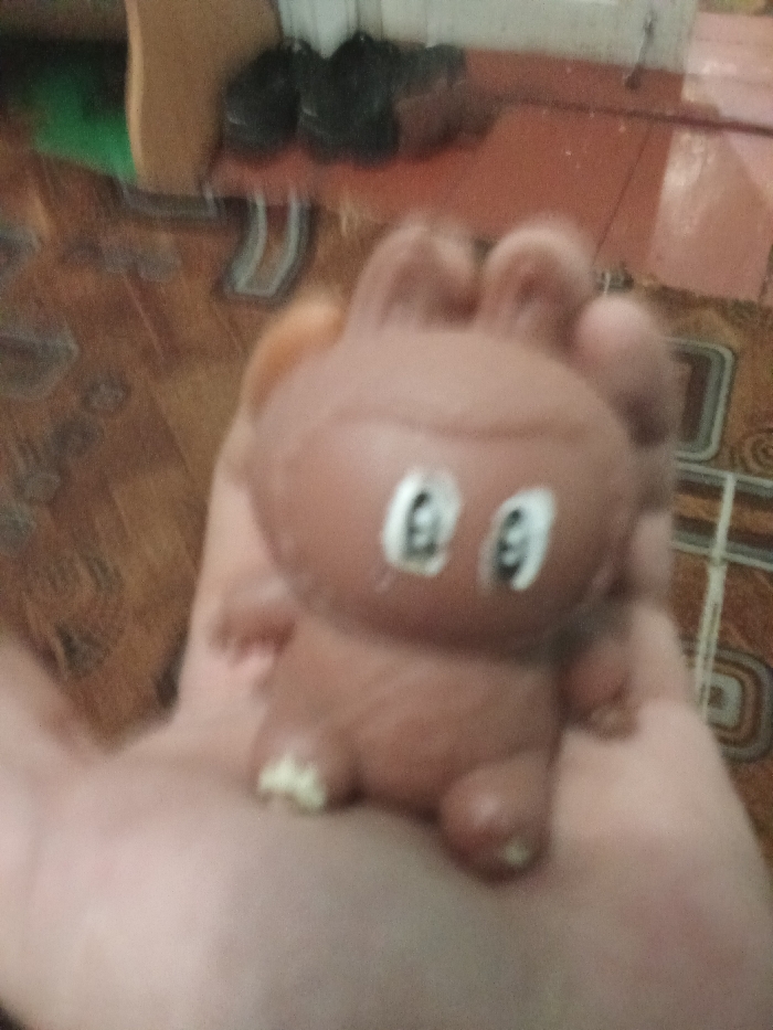 The image depicts a human hand holding a small, brown, anthropomorphic or creature-like toy. The toy is made of a soft, flexible material, featuring a rounded head with two distinct white oval eyes with dark pupils, small protrusions on its head resembling ears or tufts, and small limbs. A lighter, possibly yellowish, detail is visible on the toy's lower front. The hand, supporting the toy in an open palm, has light-toned skin. The background is out of focus, revealing an indoor environment in Yarun, Ukraine. A tiled floor with a geometric pattern in shades of brown and beige is visible. In the upper left background, dark, blurred objects, possibly shoes, rest on the floor or a low surface, alongside a vague green element. The scene is illuminated by ambient indoor light.