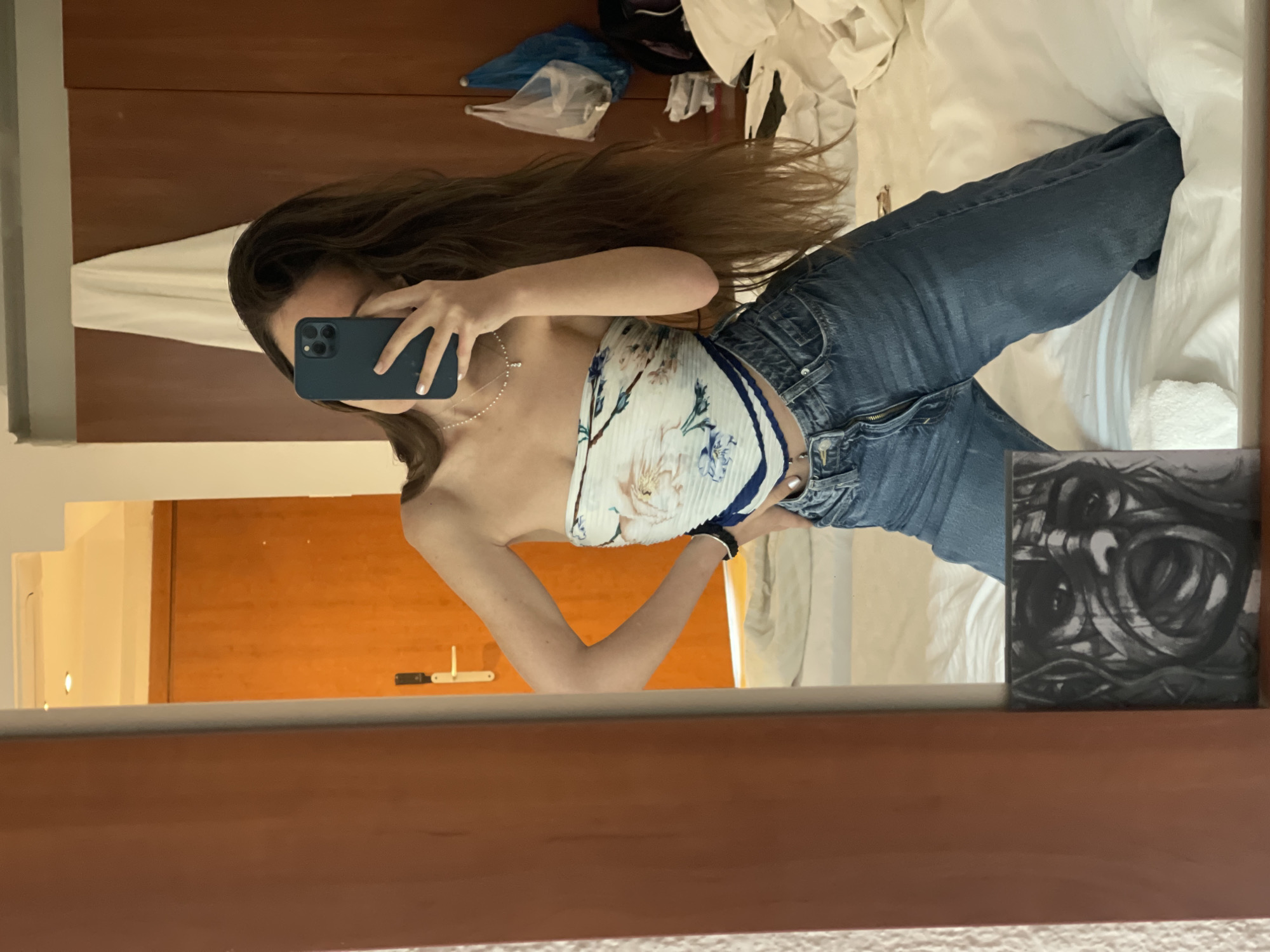 A woman is taking a selfie in a mirror. She has long brown hair and is wearing a floral scarf top and blue jeans. She is holding a dark blue smartphone. The background shows a bed with white linens, a brown wooden headboard, and a piece of artwork in the lower right corner. The reflection also shows a wooden door with a latch. No specific location within the Nea Makri Municipal Unit can be determined from this image.