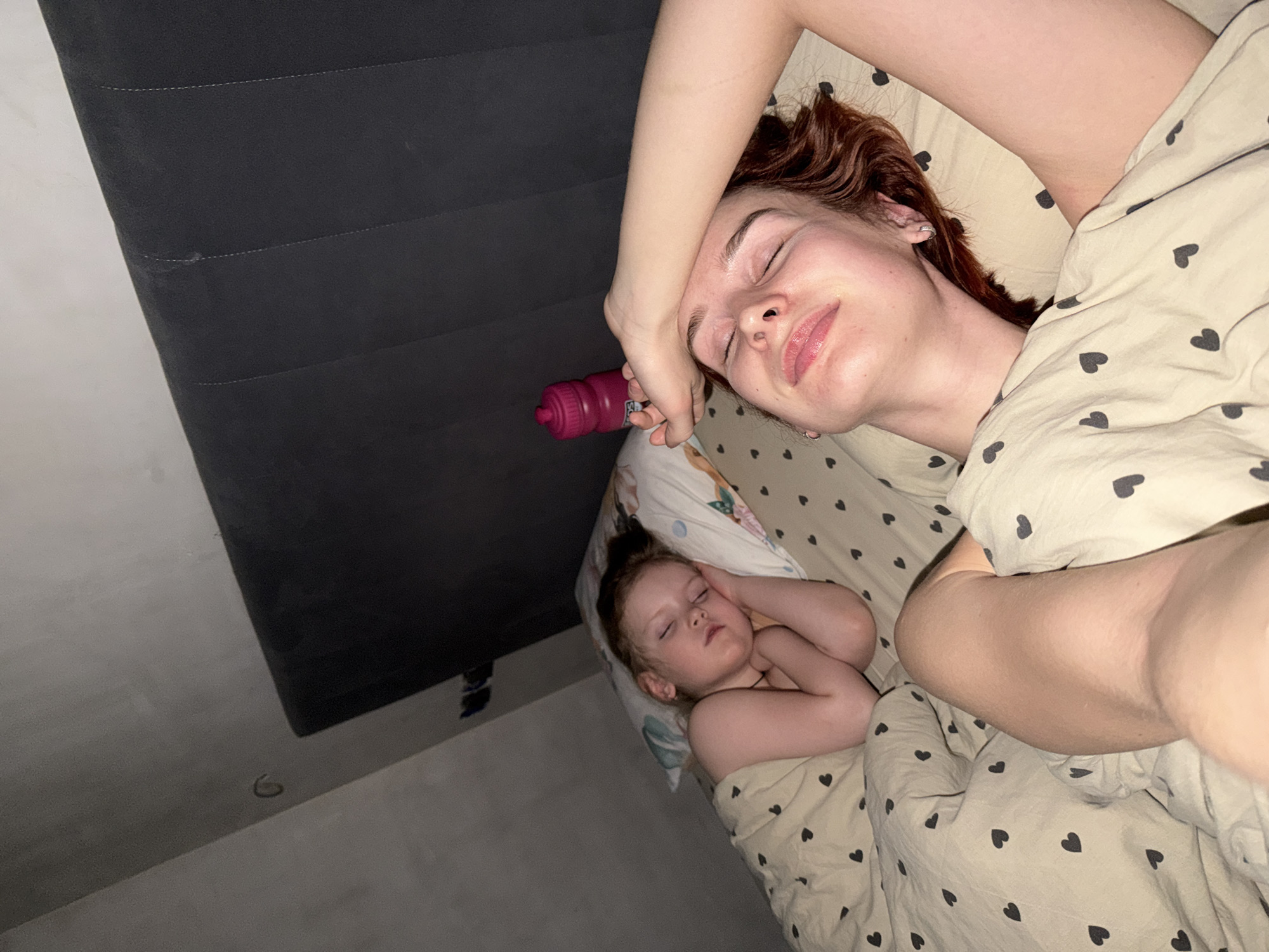 This image depicts an indoor scene within a bedroom, identified as being located in Chaikovsky, Russia. Two individuals, an adult female and a young child, are observed in a bed, both appearing to be asleep.
The adult female, positioned in the foreground and to the right, lies on her back with her eyes closed. Her left arm is bent, with her hand resting on her forehead. She has reddish-brown hair and is partially covered by a light-colored duvet patterned with small dark hearts. Her bare right arm is visible extending upwards.
To the left and slightly behind the adult, a young child lies on their left side, facing the adult. The child also has eyes closed, suggesting sleep. Their arms are bent with hands near their chest. They are covered by the same heart-patterned duvet. The child's head rests on a pillow with a colorful, abstract, or floral pattern.
Between the adult's head and the headboard, a fuchsia-colored water bottle with a textured surface is visible. The bed features a dark grey upholstered headboard with vertical stitching. The bedsheets and duvet share the light background with dark heart patterns. The room's background consists of a light grey wall, possibly part of a sloped ceiling or attic space, with a dark corner section above the child. A small, dark, curved object is affixed to the light grey wall in the upper left.
