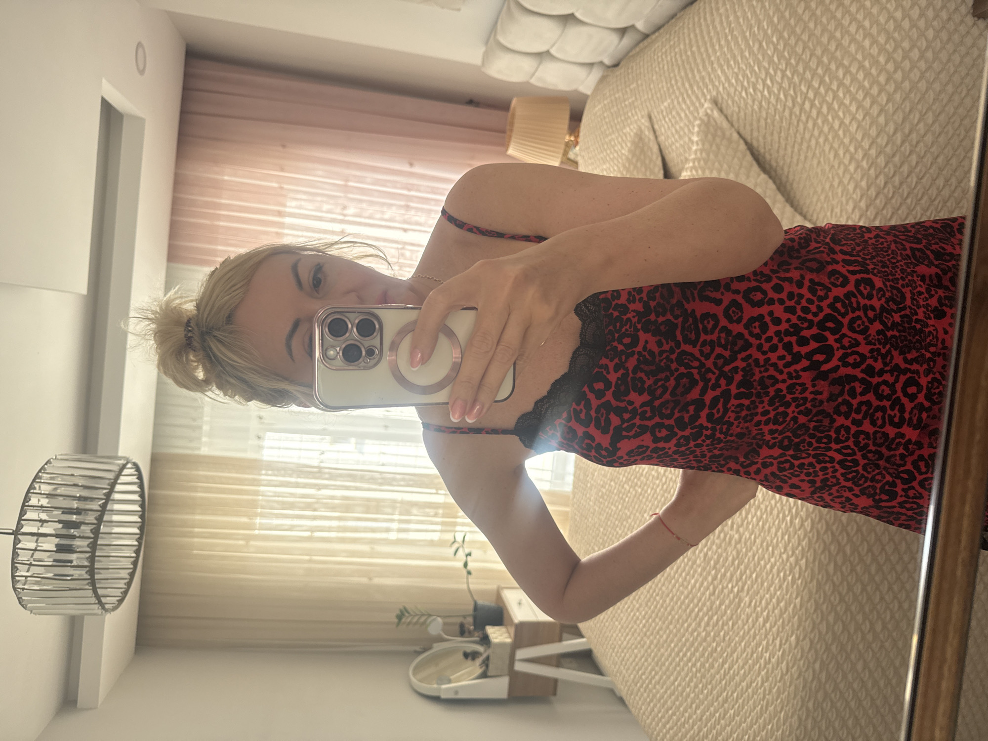 A woman is captured taking a mirror selfie in what appears to be a bedroom. She is seen from the chest up, holding a light-colored smartphone with a multi-lens camera system and a visible magnetic ring accessory, which partially obscures her face. She has blonde hair, possibly tied back, and is wearing a red and black leopard print camisole or slip dress with black lace trim. A delicate necklace is visible, and a small red string bracelet adorns her wrist. Her nails are manicured with a French tip style.
The background, reflected in the mirror, reveals a well-appointed bedroom. To the woman's right, a bed with a light-colored, textured headboard and quilted bedding is prominent. A lamp with a pleated shade is visible on a bedside table. To her left, a modern, cylindrical light fixture with clear crystalline elements hangs. Soft, natural light enters the room through sheer, light-colored curtains. Another bedside table with a small potted plant and other decorative items is also visible. The walls are white, suggesting a clean and bright interior space. The overall impression is a casual, everyday activity in a private dwelling. No legible text is present in the image.