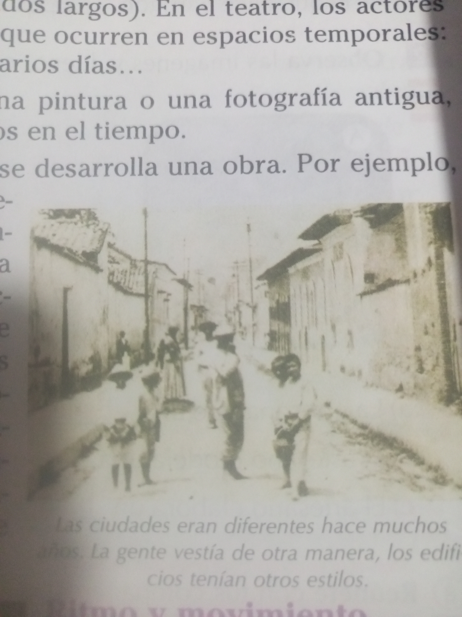 The image is a black and white photograph depicting a street scene in an old city, likely from the early 20th century. The photograph is slightly blurred and appears to be a scanned or photocopied image of a printed photograph. In the foreground, several figures, including children and adults, are walking along a dirt road lined with buildings on either side. The buildings have a rustic appearance with tiled roofs and visible architectural details that suggest an older style. One of the children in the foreground appears to be wearing a hat. The background shows more buildings and a lamppost, further emphasizing the urban setting. The image is accompanied by text in Spanish, which discusses concepts related to theater, actors, time, painting, photography, and historical context. It also mentions "La Tagua, Venezuela" in the context of cities being different many years ago and people dressing differently. However, the photograph itself does not contain any explicit visual cues that definitively identify the location as La Tagua, Venezuela. The scene suggests a historical moment where daily life is captured in a simple, unposed manner.