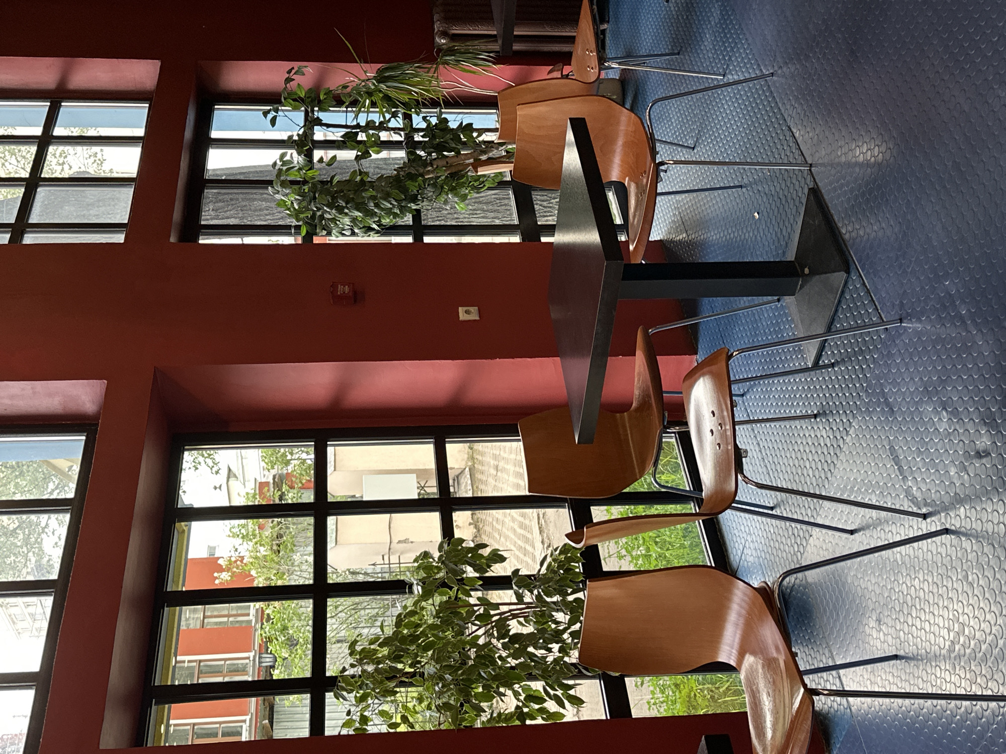The image shows the interior of a building, possibly a cafe or restaurant, located in Moscow, Russia. The walls are painted a deep red color, and large windows offer views of the outside. Through the windows, buildings typical of Moscow architecture are visible. There are plants, likely potted, positioned near the windows, adding a touch of greenery. Several chairs and a table are arranged within the space, suggesting a seating area for customers. The chairs are of a modern design, with wooden seats and metal frames. The floor is covered in a patterned rubber flooring.