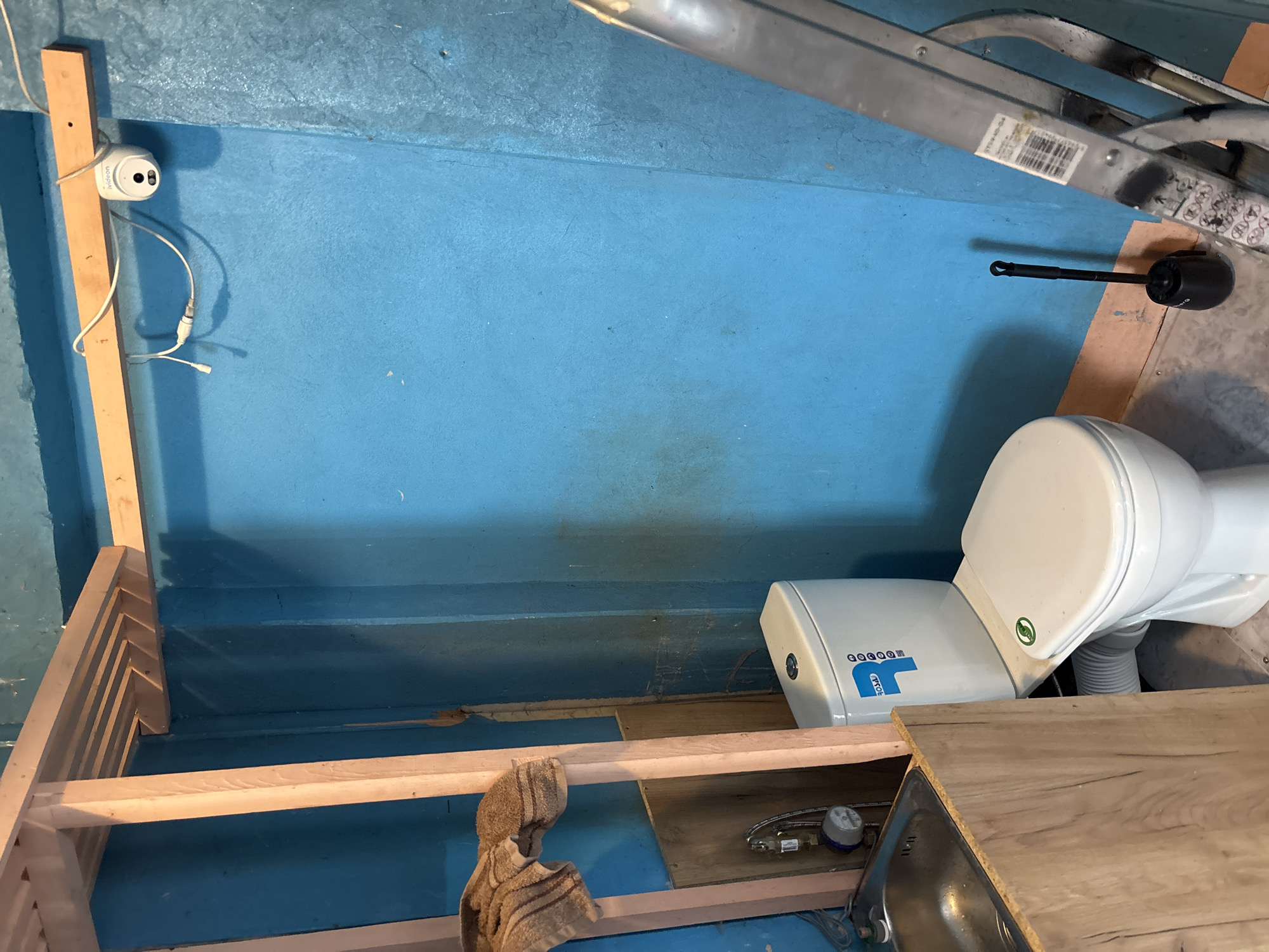 This image depicts the interior of a small, unfinished room or enclosure, likely a bathroom or a utility space. The walls and floor are painted a vibrant blue. In the foreground, a wooden structure, possibly a bench or shelving unit, is partially constructed. A beige towel hangs from the wooden frame.
To the right of the frame, a white toilet is installed. It appears to be a compact model, possibly designed for a smaller space. The toilet has a brand logo with blue lettering that reads "THEET" and a green circular sticker with a leaf design on the tank. Adjacent to the toilet, a white tank, possibly a water heater, is visible.
Further to the left, a small white security camera is mounted on a wooden post. It is connected by white wires. In the upper right quadrant of the image, a silver ladder is leaning against the blue wall. A barcode label is attached to one of the ladder's rungs.
The scene appears to be in an indoor setting, with artificial lighting. There are no people visible in the image. The overall impression is of a space undergoing renovation or construction. The specific location of Vileyka, Belarus, is noted.
