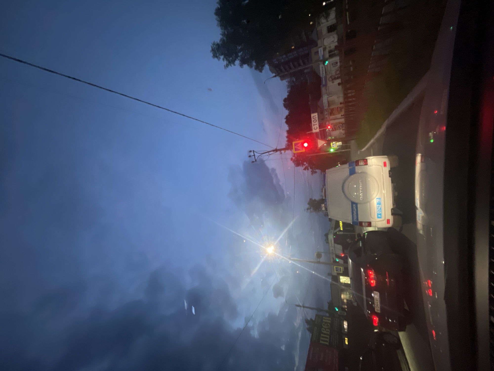 The image captures a street scene in Votkinsk, Russia, during twilight or dusk. The sky is a deep blue with dark, dramatic clouds on the left, illuminated by a bright streetlamp that creates a lens flare effect. In the foreground, the view is from inside a vehicle, showing a portion of the dashboard.
On the road, several vehicles are visible. A white police SUV with "ПОЛИЦИЯ" (Police) written on its side and the license plate "У 0742 18" is prominent, stopped at a traffic light. To its left, a dark-colored sedan with a Russian license plate "Е044ХС 18" is also stopped. Further down the road, a traffic light shows red. A stop sign ("СТОП") is visible above the traffic light.
On the left side of the road, there are buildings with illuminated signs. One sign appears to have Cyrillic text, possibly "ШВЕСИ". Trees and more buildings are visible on the right side of the street, with streetlights and building lights adding to the ambiance of the scene. Power lines crisscross the sky. The overall impression is of a typical urban environment transitioning from day to night, with police presence evident.