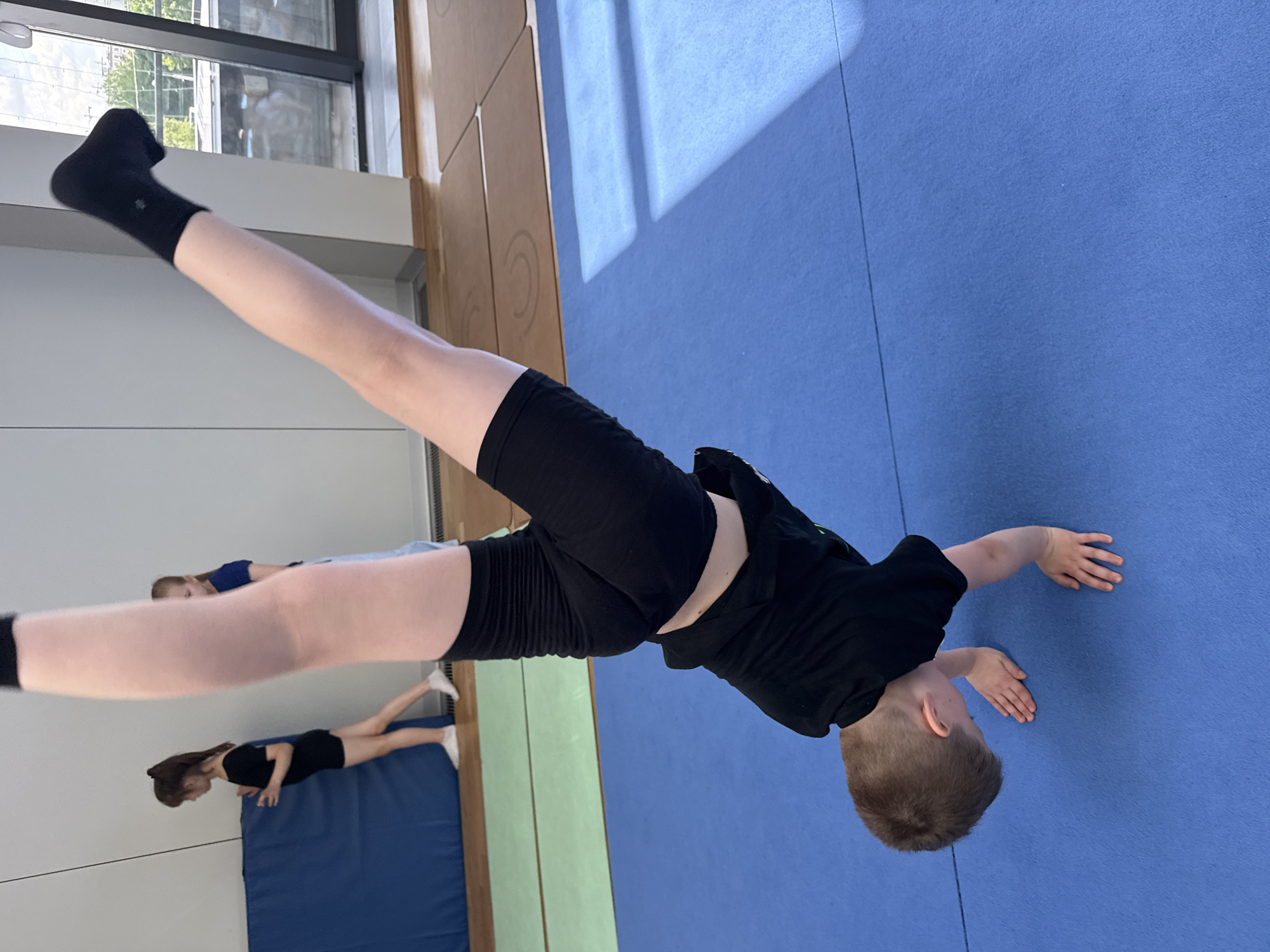 This image captures an indoor scene, likely a gymnasium or sports hall, where children are engaged in gymnastic or acrobatic training in Khimki, Russia. The main subject is a young child, appearing to be male, positioned centrally and inverted in an acrobatic pose. He is wearing black athletic shorts, a black t-shirt, and black ankle socks, one of which clearly displays the brand "ALO". His hands are placed on the blue floor mat, and his legs are extended straight upwards, indicative of a handstand or similar inverted movement. His head, with short brown hair, is visible near his hands, looking towards the floor.
In the background, two other children are visible, further away. One child, wearing a black top and shorts, is lying or kneeling on a separate blue mat. Another child in a blue top is also present, partially obscured, suggesting they are participating or observing the activity.
The setting features a prominent blue floor mat covering a large area. Adjacent to this is a section of light brown wooden flooring with subtle circular patterns. A segment of a light green wall is visible in the mid-ground. Large windows line an upper wall, allowing significant natural light to enter, creating sharp, diagonal shadows on the blue mat, indicating it is a bright, sunny day. The overall scene depicts a focused and active training environment. Apart from "ALO" on the sock, no other visible text is discernible.