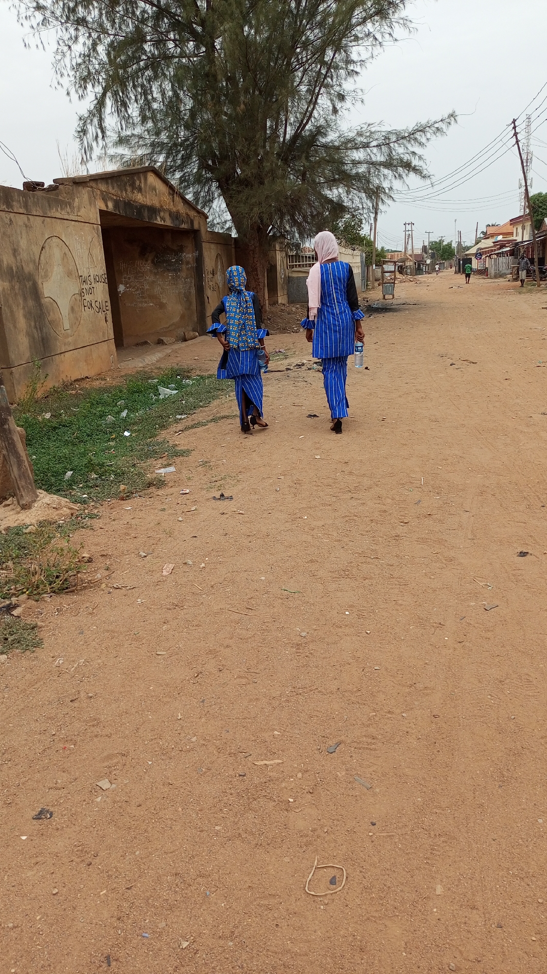 Two women are walking down a dirt road in Sabon Tasha, Nigeria. The woman on the right is wearing a pink hijab, a black shirt, and a blue and white striped outfit, and is carrying a water bottle. The woman on the left is wearing a blue and white striped outfit with a matching headscarf. They are walking away from the viewer. In the background, there is a building with graffiti on the wall that reads "THIS HOUSE IS NOT FOR SALE." There is a large tree between the building and the women. On the right side of the road, there are other buildings and people further down the road. Overhead, there are electrical lines. The ground is a reddish-brown color and there is some trash scattered along the roadside.