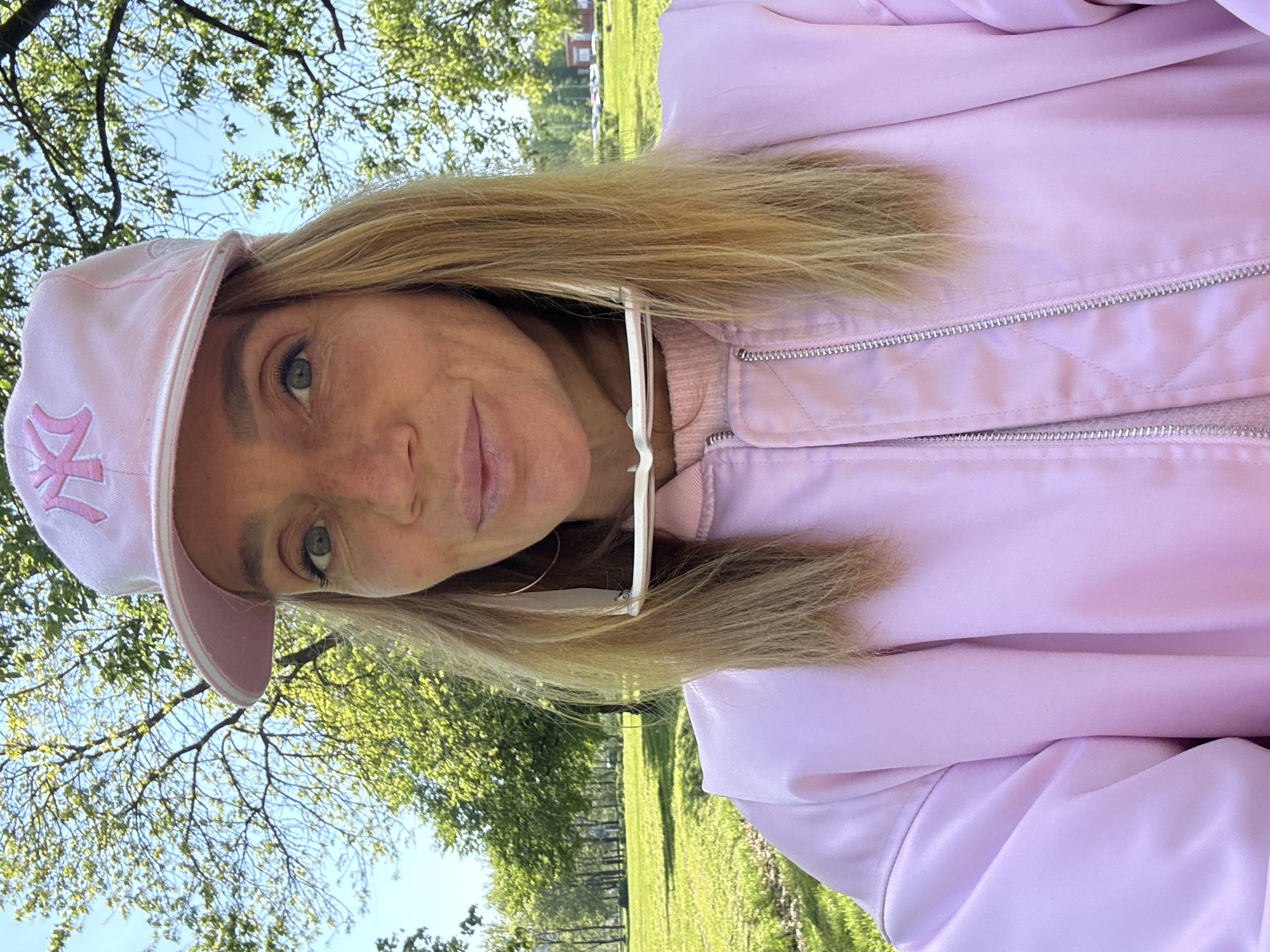A woman is centered in a selfie shot outdoors. She wears a light pink jacket and a matching baseball cap with a "NY" logo. A pair of white sunglasses are casually resting on her chest. She has blonde hair, and her eyes are visible. The background reveals a green grassy area with trees, suggesting a park or garden setting. In the distance, a building is visible. Based on the trees and building structure, the location appears to be a public park in Saint Petersburg, Russia.