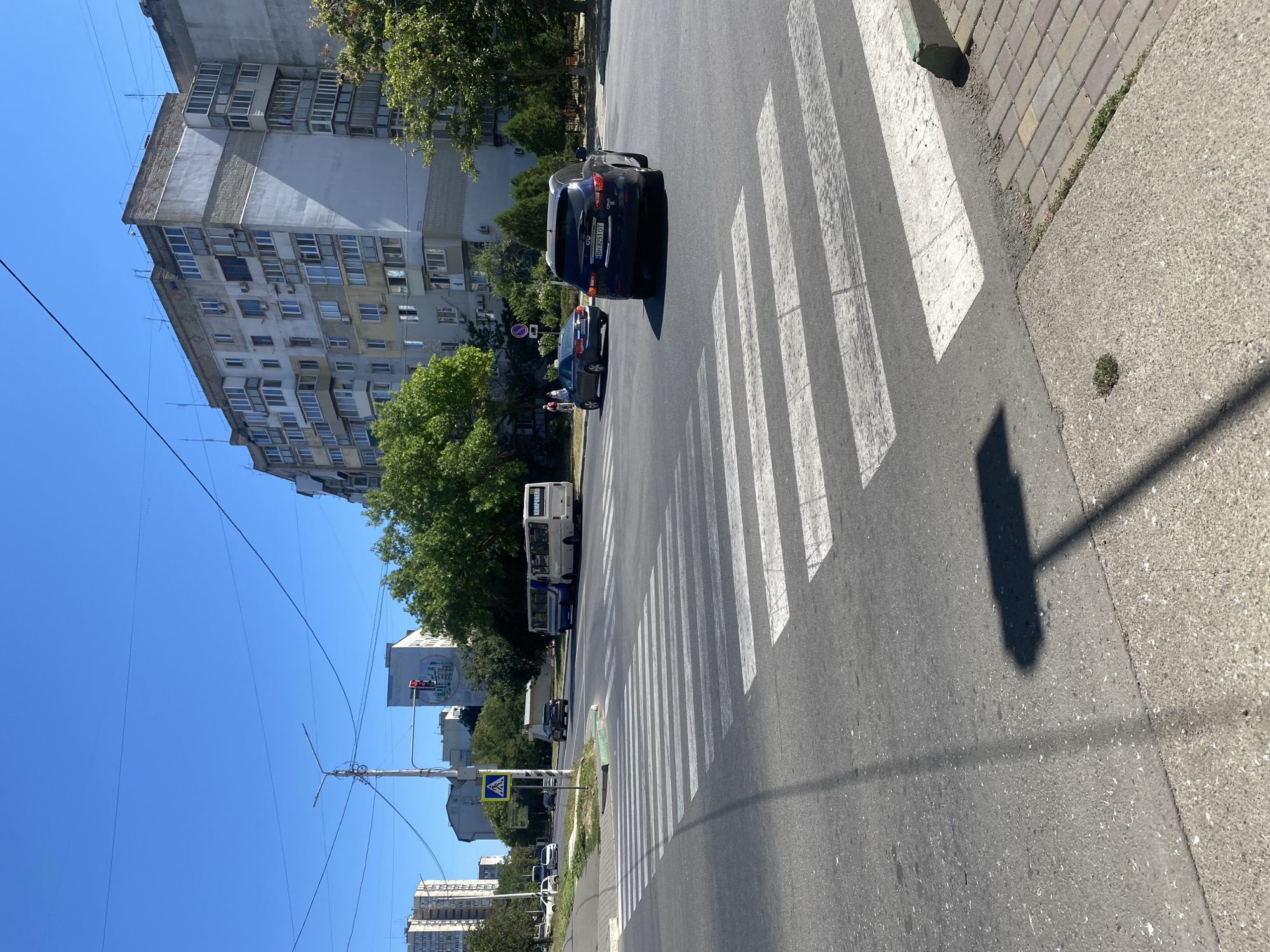 A street scene in Chornomorsk, Ukraine, features a pedestrian crossing with a blue sky overhead. Several cars are visible, including a dark blue Infiniti SUV with license plate BH 8251 OT, a dark sedan, and a white car. A white bus with the word "KOMPONENE" written on its rear is also present, as is a blue and white bus. Two pedestrians are walking on the sidewalk near the buses. The environment includes apartment buildings and trees. A traffic light is visible, currently red. A blue and white pedestrian crossing sign is posted on a pole. Shadows of streetlights and a pedestrian crossing pole are cast across the road and sidewalk. The weather appears sunny and warm.