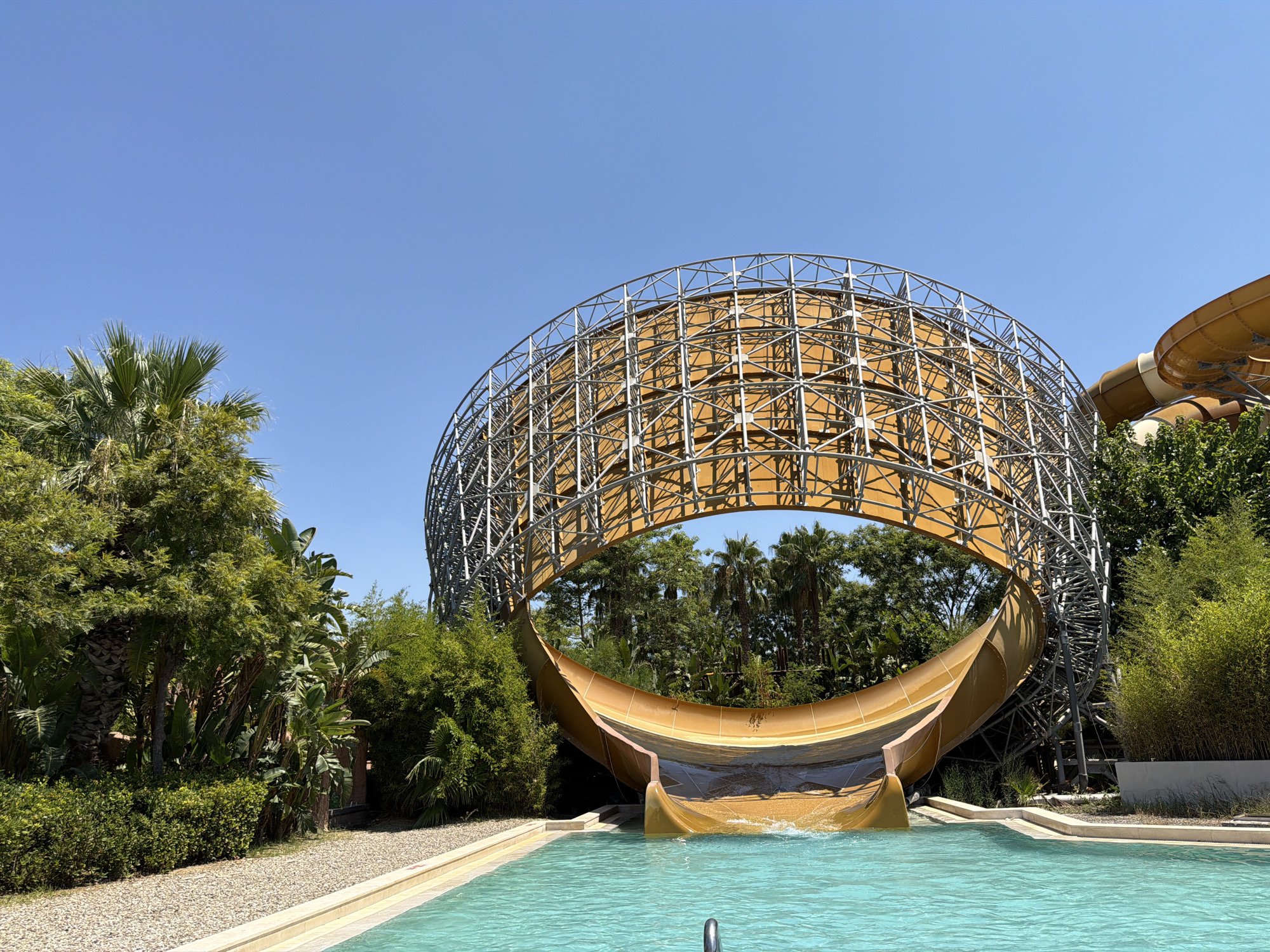 A wide shot shows a large, circular water slide structure at a water park. The slide is yellow and appears to be made of plastic, with a metal scaffolding supporting it. The slide empties into a bright blue swimming pool. Lush green trees and plants surround the area, and the sky is clear and blue. In the background, another yellow water slide can be seen. There are no people in the image.