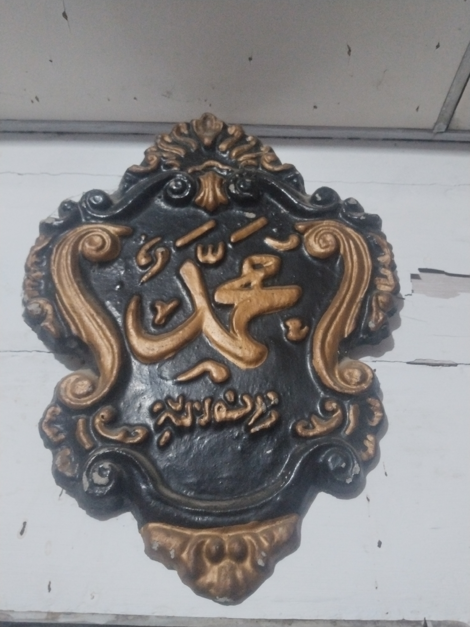 A decorative plaque, predominantly black with intricate gold detailing, is affixed to a light-colored wall, likely indoors. The plaque features prominent Arabic calligraphy rendered in gold relief. The main text clearly spells "Muhammad" (محمد). Below this, a smaller line of Arabic script is visible, which typically accompanies the name of the Prophet Muhammad, often a blessing or a title like "Messenger of God." The plaque's frame is highly ornate, characterized by elaborate gold scrollwork and volumetric decorative elements, suggesting a traditional Islamic or Baroque aesthetic. The background wall is off-white and shows signs of wear, including visible cracks and some texture. A section of what appears to be a ceiling or upper wall structure, possibly a panel or beam, is visible at the top. The lighting is diffused, and no specific time of day or outdoor weather conditions are discernible. The object serves as a cultural or religious display, consistent with the context of Lhokseumawe, Indonesia.