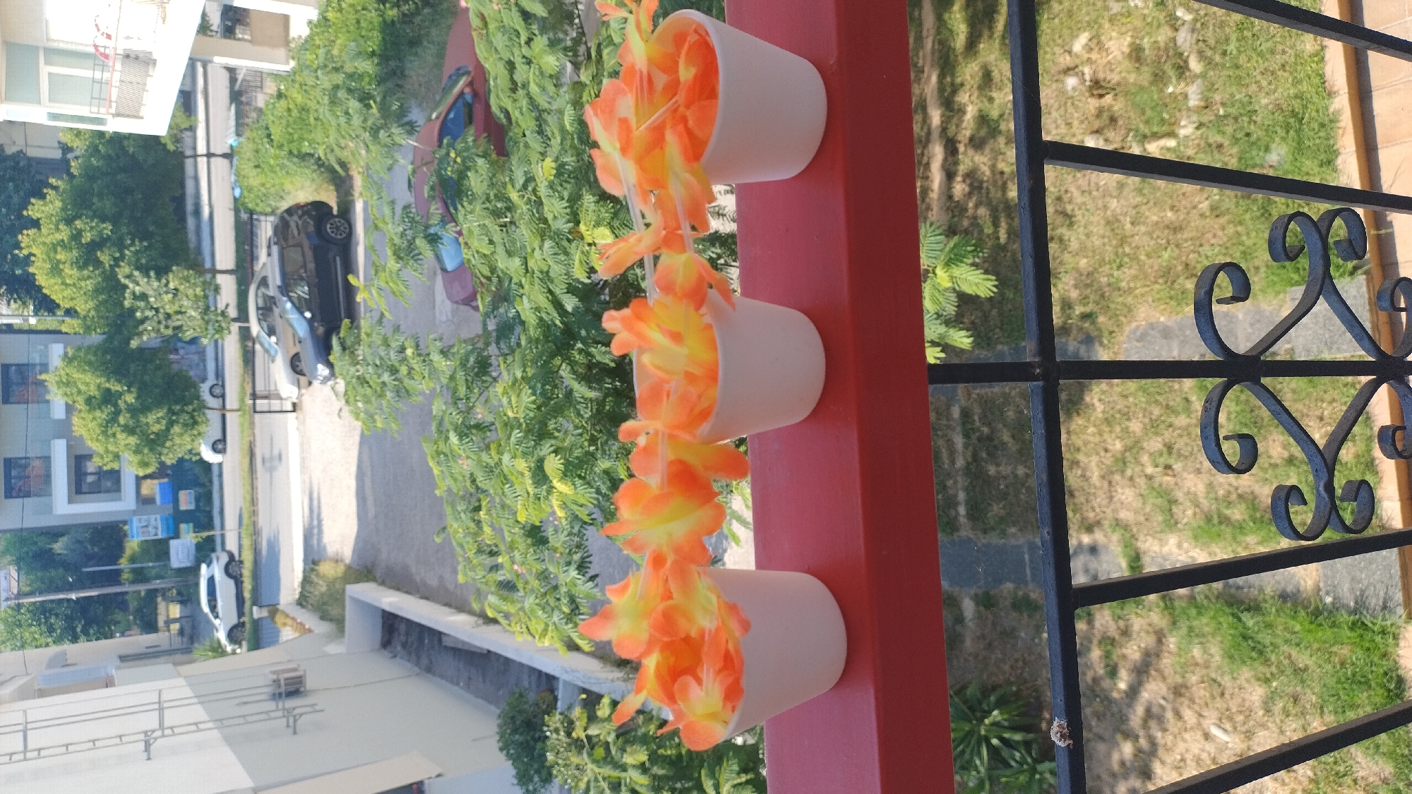 This image shows a close-up of three white pots filled with orange and yellow artificial flowers, arranged on a red railing. In the background, there is a blurred view of a street with cars, buildings, and lush green trees, suggesting an outdoor urban environment. The overall scene appears to be on a balcony or elevated platform overlooking the street. The Perea, Greece location is inferred from the context provided, but not directly visible in the image.