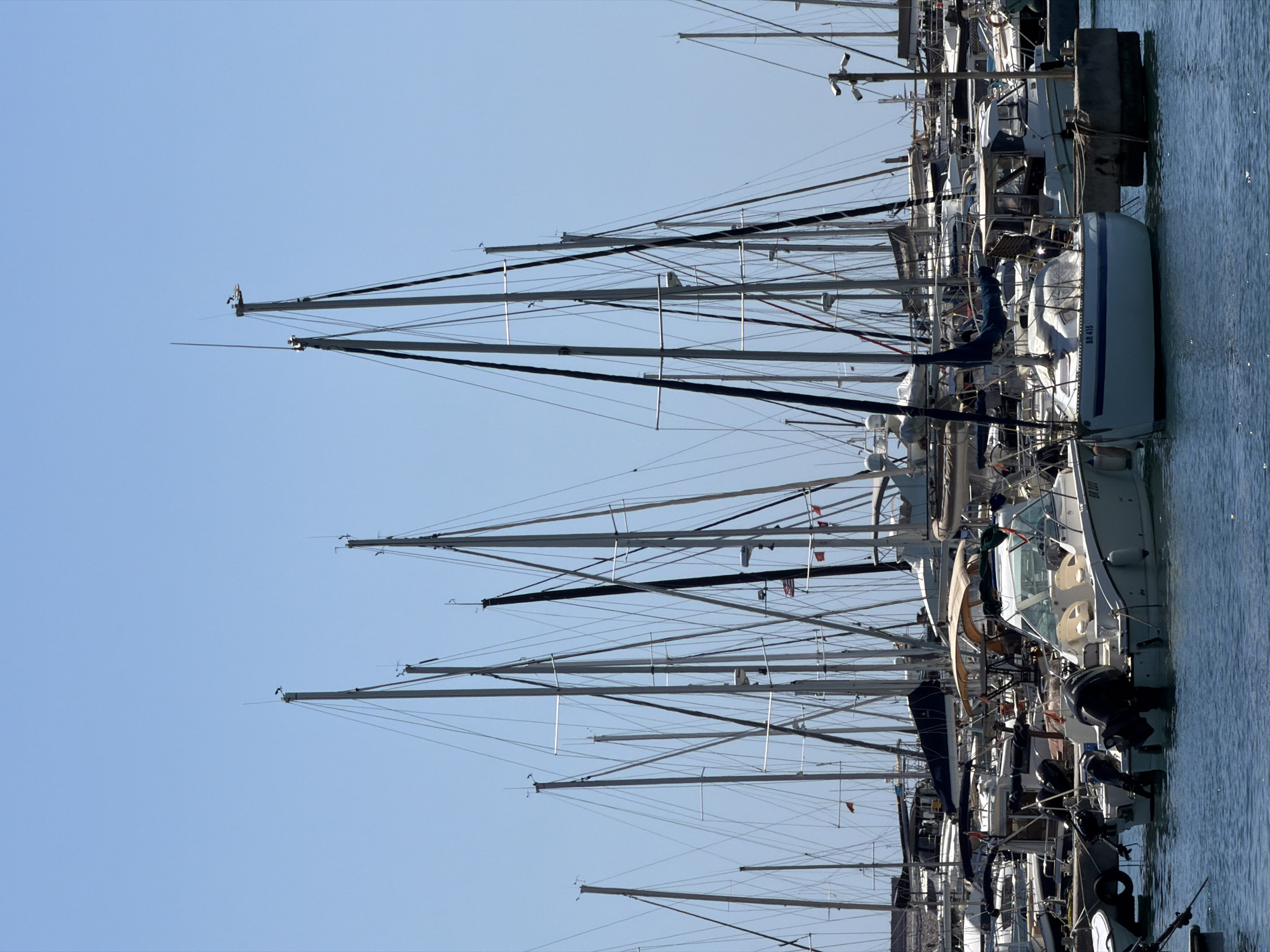 The image depicts a marina filled with numerous sailboats and smaller motorboats docked under a clear blue sky. The foreground features a white motorboat with dark outboard engines. Several sailboats with tall masts dominate the middle ground, their rigging clearly visible against the sky. In the background, more boats are packed closely together, and a few flags can be seen flying. The water is a deep blue, and the overall scene suggests a sunny day in a busy harbor. This scene is located in Šušanj, Montenegro.