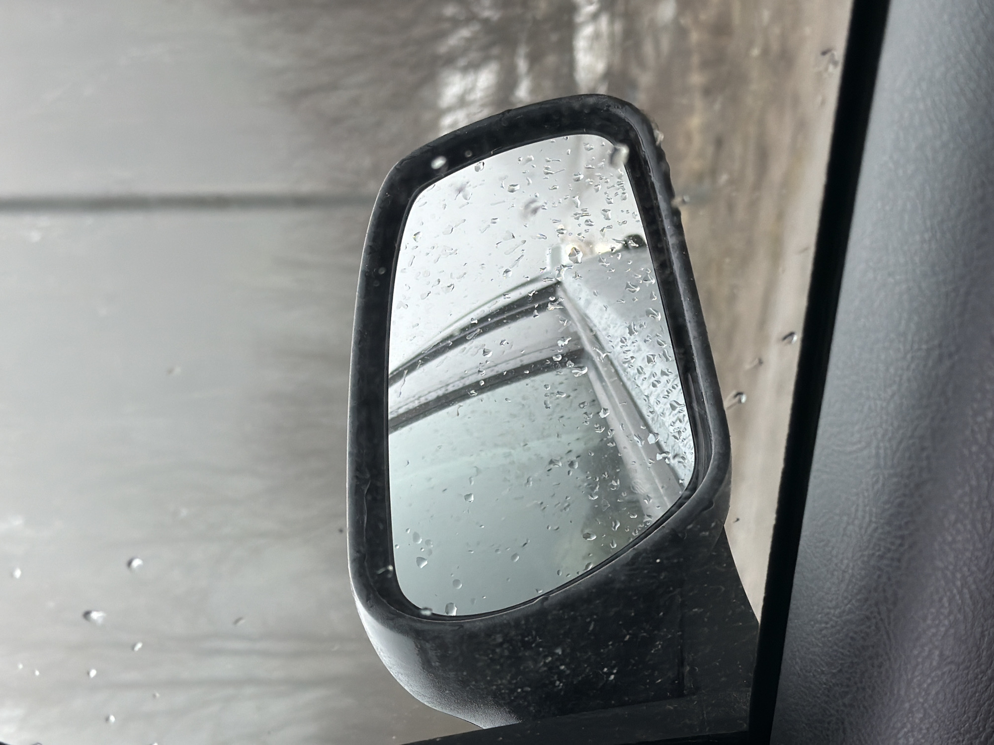 The image depicts a side view mirror of a vehicle, covered in raindrops. The mirror reflects a blurred image of another vehicle, possibly driving behind. Outside the car window, the scene appears to be overcast and wet, possibly in an urban setting. The interior of the car is visible on the right side of the frame. Location data of Chaikovsky, Russia is not possible to determine from the image alone.