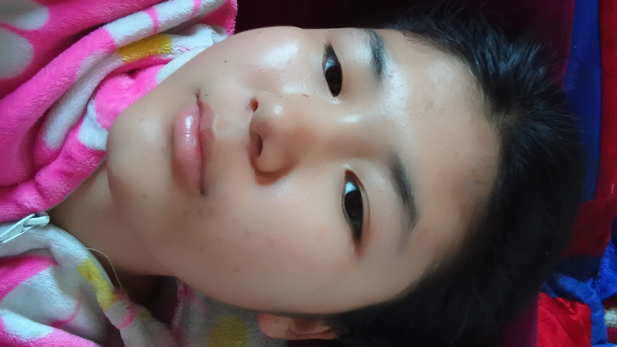 A close-up selfie shows a young Asian person lying down. Their face is angled slightly towards the viewer, with their eyes open and looking forward. They have dark hair and smooth skin. They are wearing a pink hooded jacket with white and yellow accents. The background is blurred and appears to be a red and blue fabric. The scene is indoors, likely a bedroom, given the soft furnishings. The image's context as being taken in Uchkun, Kyrgyzstan, is not visually discernible.