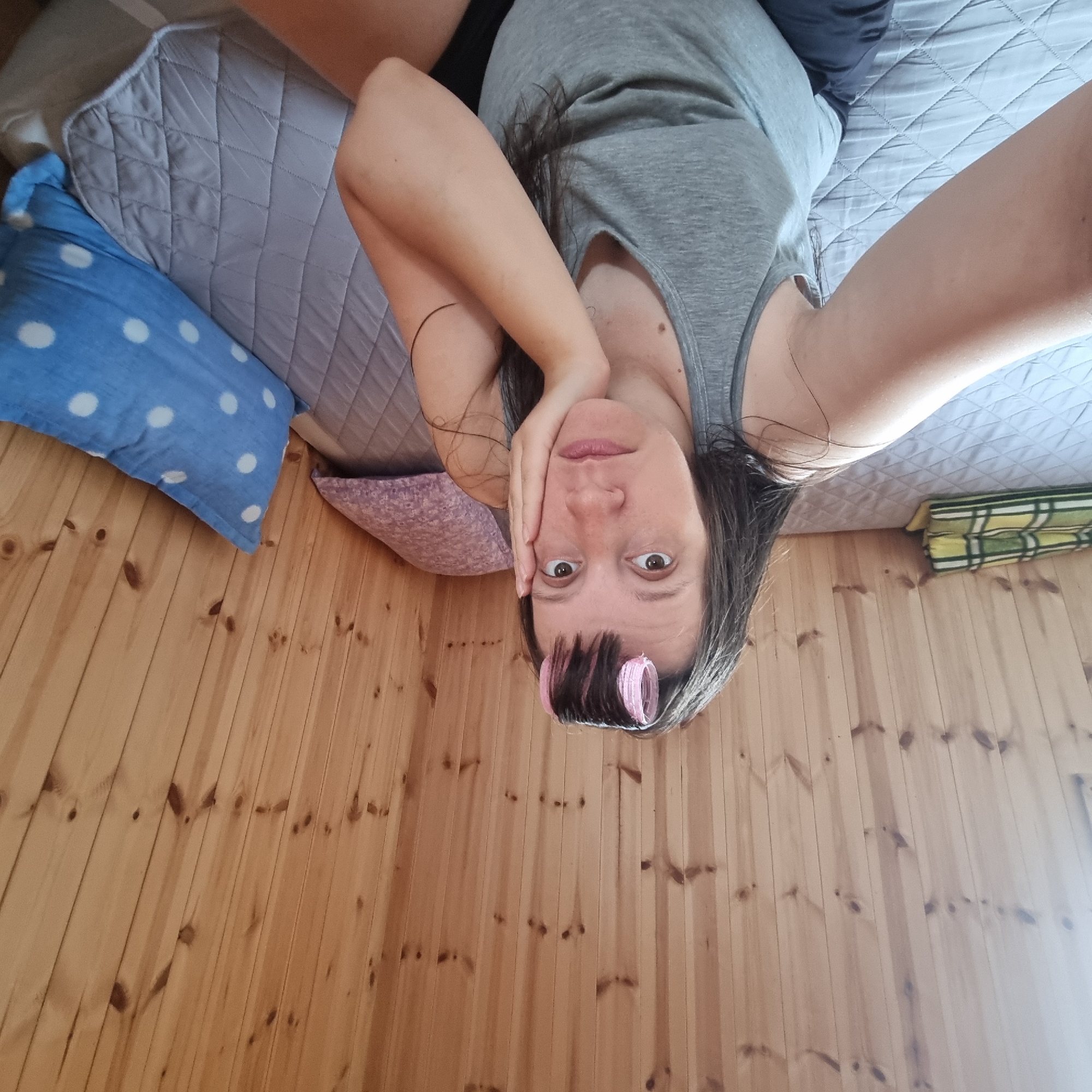 A woman with dark hair and pink hair rollers in her bangs is sitting on a bed and looking at the camera with a somber expression. She is resting her right cheek on her hand. She is wearing a grey tank top and dark shorts. The bed is covered with a grey quilted mat and there are two pillows on it, one pink and one blue with white polka dots. The background is a wooden paneled wall. The scene appears to be in a rustic or cabin-like setting. This image was taken in Dunnae-myeon, South Korea.