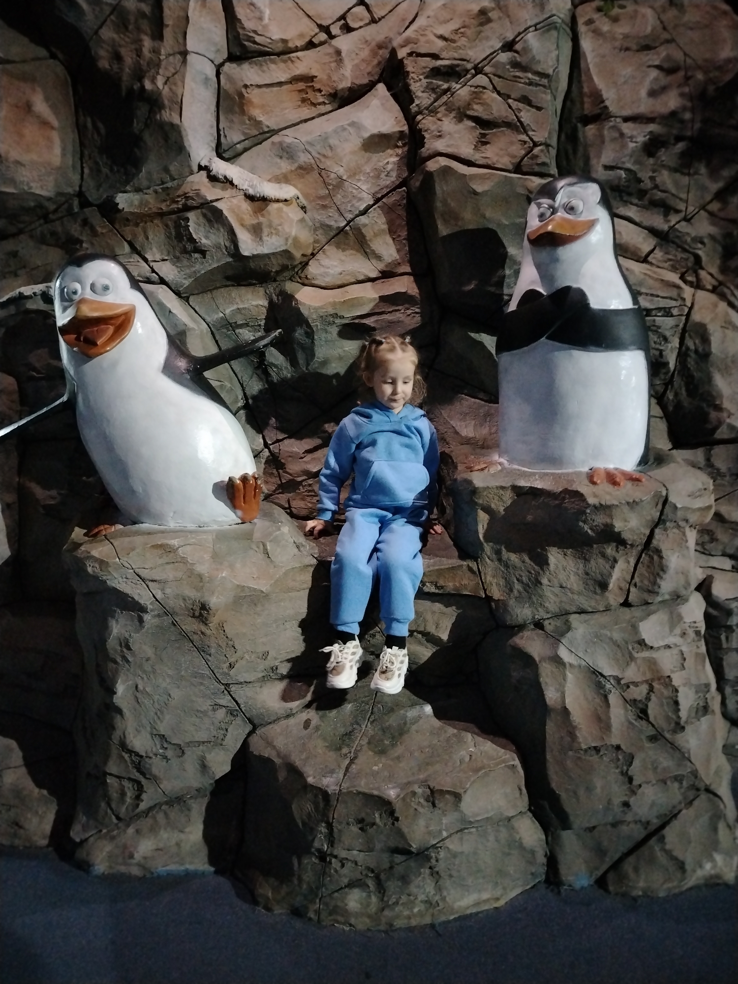 A young child sits on a rock formation between two cartoon penguin statues. The child is wearing a light blue hooded sweatshirt and pants and white sneakers with black socks. The penguin to the left is smiling with its wings slightly outstretched, while the penguin on the right has its arms crossed and looks displeased. The background is a faux rock wall. The scene is taken at the Kazan Oceanarium in Kazan, Russia.