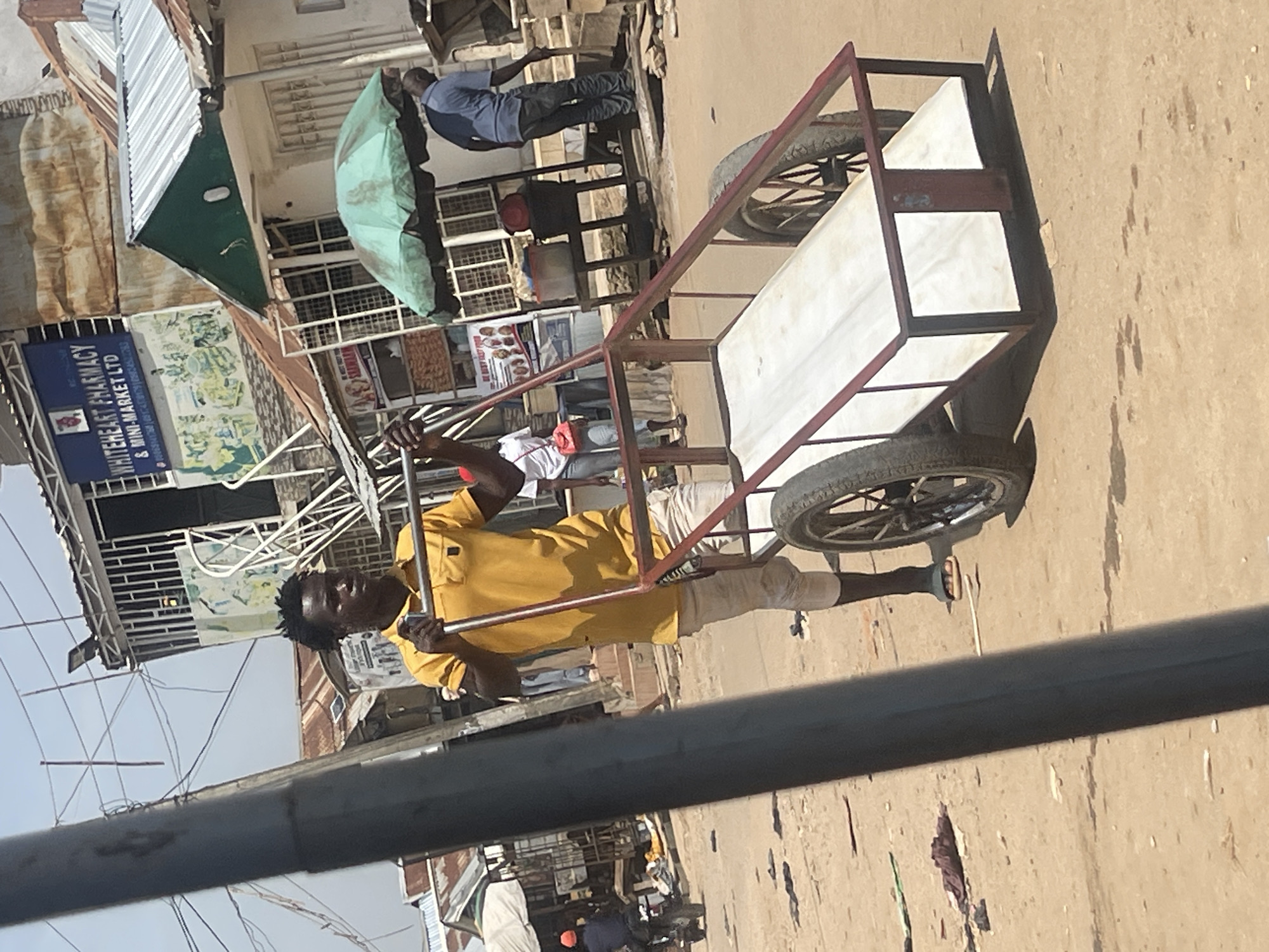 In Abuja, Nigeria, on a dusty, unpaved street, a young man with dark, spiky hair and a determined expression pushes a red metal cart with two large spoked wheels. He wears a bright yellow polo shirt and light-colored, knee-length trousers. The cart has a white, padded bed and a red frame. In the background, buildings with corrugated metal roofs line the street. A green umbrella shades a vendor's stall. Further down the street, a man in a blue shirt and dark pants stands by a table laden with goods. A motorcycle rider with a red helmet is visible in the distance, heading away from the viewer. The ground is littered with scattered debris and shadows.