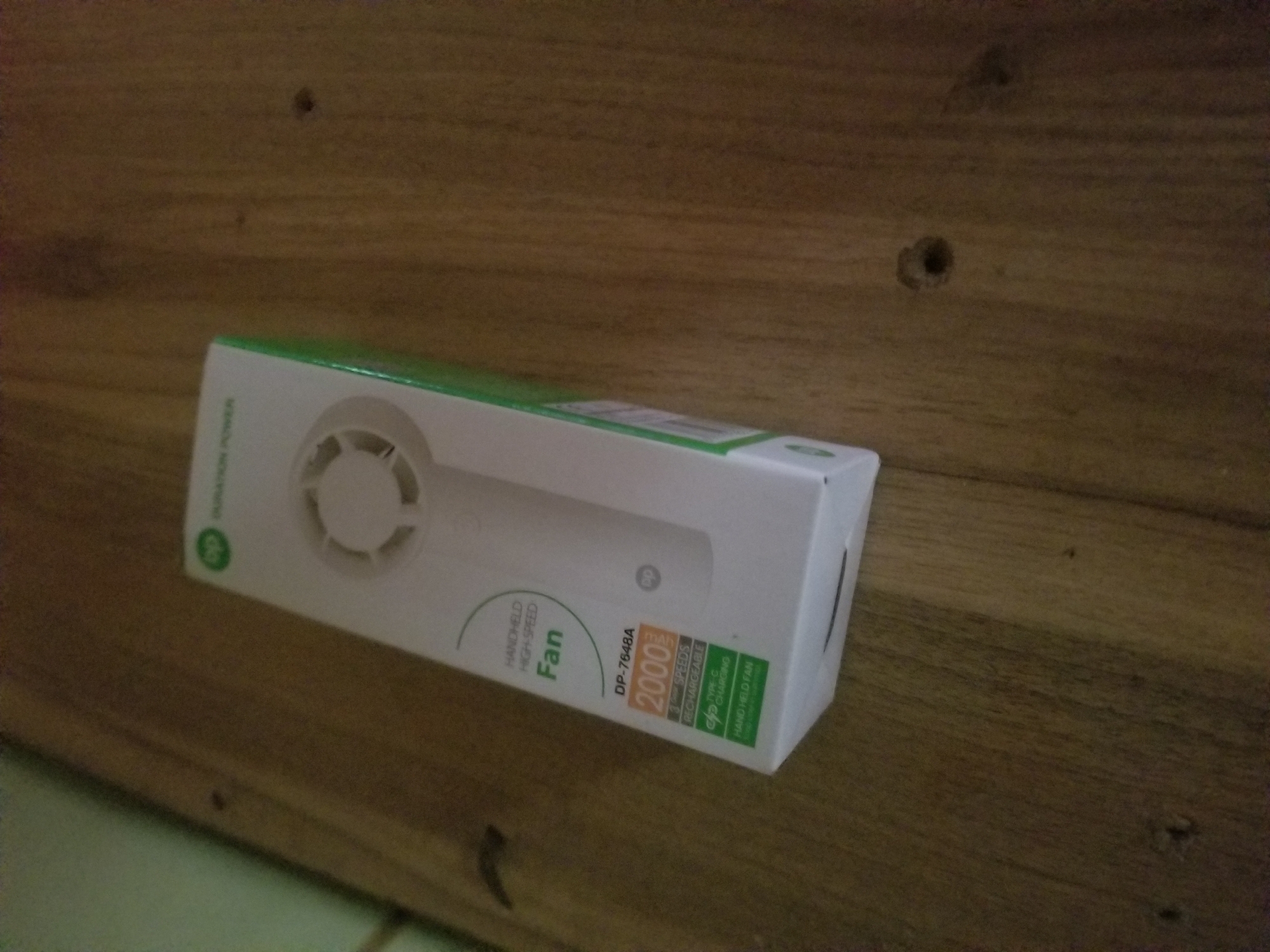 A white and green box for a handheld fan sits on a wooden surface. The box has text and an image of a fan on it. Visible text includes "HANDHELD HIGH-SPEED Fan", "DP-7648A", "2000mAh", "3 SPEEDS RECHARGEABLE", and "TYPE C CHARGING HAND HELD FAN". The wooden surface appears to be a table or shelf with visible grain and a few small holes. The lighting is dim, suggesting an indoor setting. There are no people or animals visible in the image.