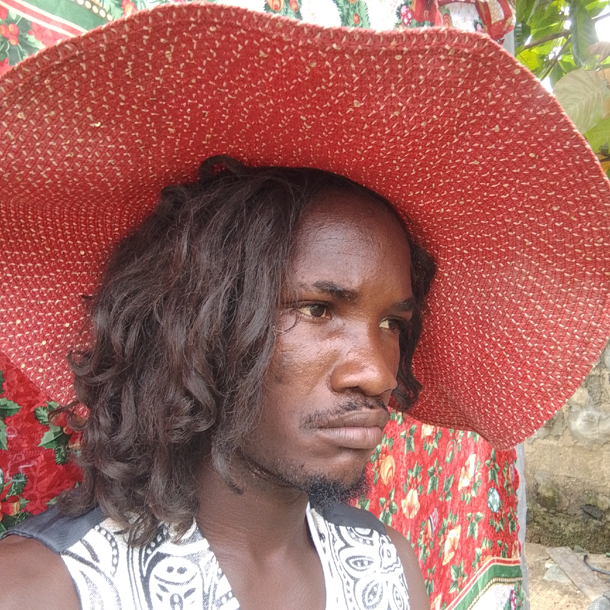 The image shows a close-up, head-and-shoulders portrait of a dark-skinned individual wearing a wide-brimmed red hat adorned with a textured pattern. The hat covers most of the person's head, with dark, wavy hair visible around the face and shoulders. The individual is looking to the right, with their face partially obscured by the hat. They are wearing a white garment with a black and white patterned design. The background is a colorful fabric with a red base and a floral pattern in green, yellow, and white. A stone wall and some greenery are visible to the right of the frame. The lighting suggests an outdoor setting. This image is not from Ado, Nigeria.