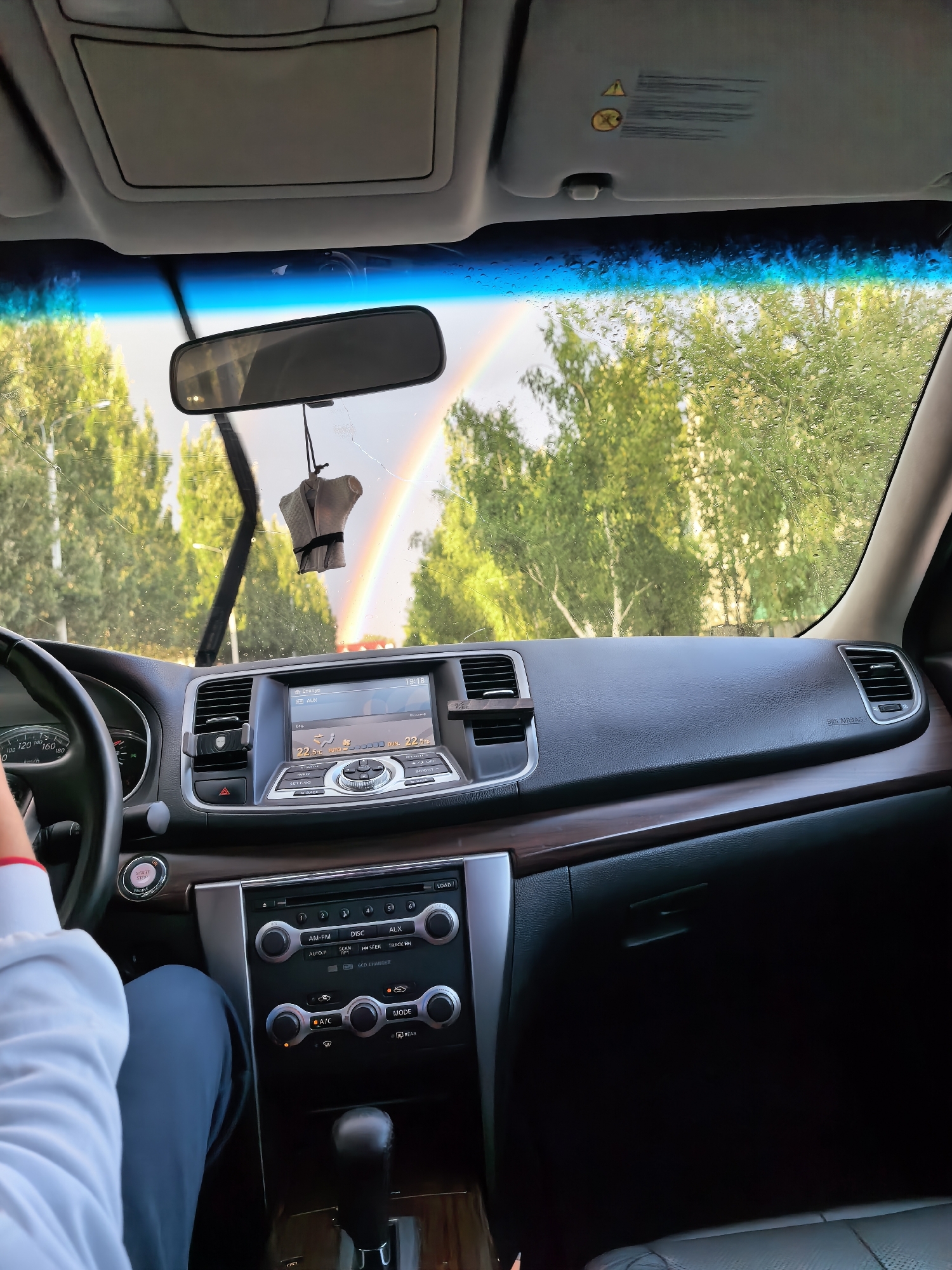 The image shows the interior of a car, looking out through the windshield at a rainbow. The dashboard of the car is visible, with a central infotainment screen, climate controls, and a gear shift. A person's arm and shoulder are visible on the left side of the frame, suggesting they are the driver or a passenger. The windshield has water droplets on it and the wiper blades are visible, indicating it may have rained recently. Trees are visible outside the car, and the sky suggests it is daytime. The image is a snapshot from inside a vehicle, capturing a moment with a rainbow as a backdrop. There is no information in the image to suggest a location in Sterlitamak, Russia.