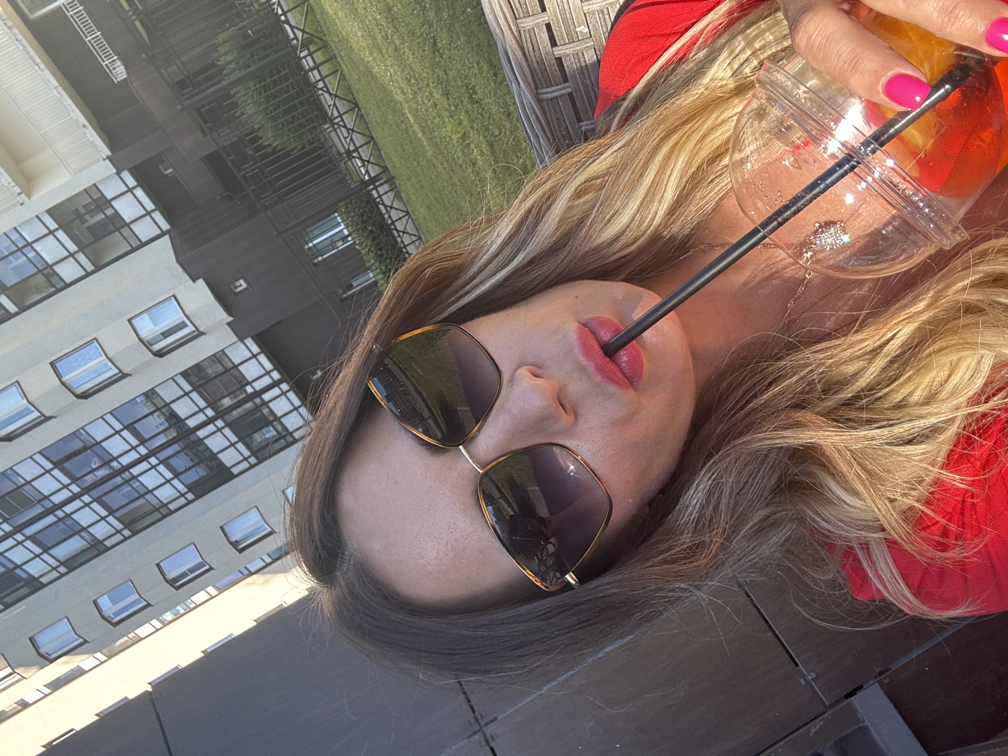A woman is taking a selfie outdoors in Saint Petersburg, Russia. She is wearing sunglasses and a red top, and holding a drink with a straw. Behind her, a modern apartment building and a grassy area with a fence are visible. The sun is shining, casting shadows and highlighting the scene.