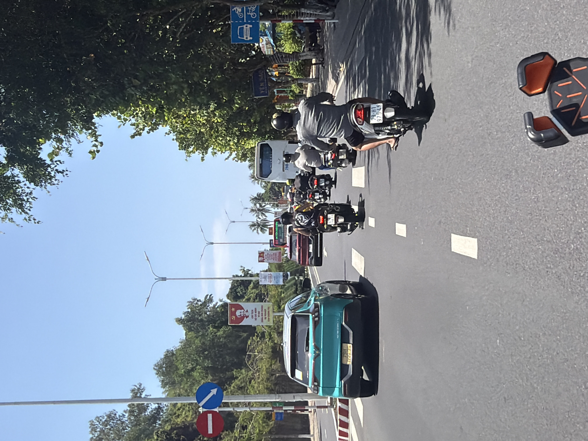 The image depicts a vibrant street scene on a sunny day in Bac Nha Trang Ward, Vietnam. The perspective is from slightly above, looking down a multi-lane road flanked by lush green trees under a clear blue sky.
The primary subjects are various vehicles navigating the road. In the foreground and midground, a stream of motorcycles and scooters, common in Vietnam, occupies multiple lanes. The closest visible motorcycle, driven by a person wearing a helmet and a grey shirt, has a license plate "79-H1 975.45". To the right, a striking turquoise-colored car is also present, featuring a license plate that reads "59C 661.85". Further down the road in the left lane, a white passenger van or minibus is visible. Streetlights, utility poles, and road signs are interspersed along the road. On the right side, partially out of frame, parts of what appears to be a black and orange protective gear item or similar object are seen on the asphalt.
The activity taking place is typical urban traffic flow. Vehicles are moving along the road, suggesting a busy thoroughfare. The clear sky and strong shadows cast by trees and vehicles indicate it is daytime, likely midday or early afternoon.
Notable details include several road signs. Near the trees on the left, a blue rectangular sign with a white bicycle icon and a bus icon indicates specific lane usage. Further down on the left, a blue circular sign with a white arrow pointing straight ahead (indicating 'straight only' or 'ahead') is paired with a red circular sign with a white horizontal bar (a 'do not enter' or 'no entry' sign). In the background, banners and signs are affixed to poles; one prominent red banner features a portrait, likely of Ho Chi Minh, with Vietnamese text below it. Another sign appears to read "YEN SAO" and "CONG TY TNHH" (Limited Liability Company). The overall setting reflects a bustling urban environment characteristic of Southeast Asia.
