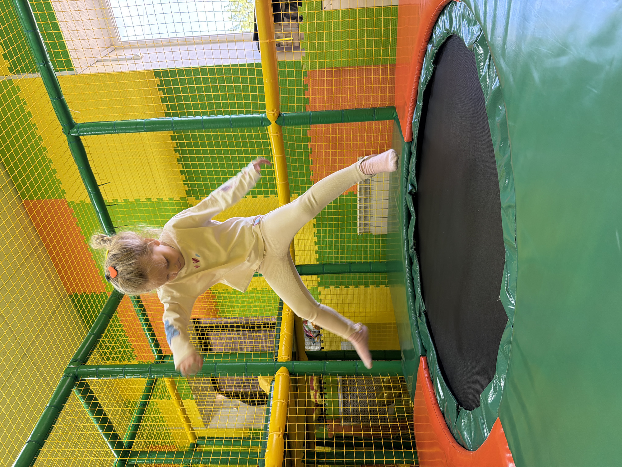 A young girl is jumping on a trampoline indoors. The trampoline has a black jumping surface and is surrounded by green and orange padding. The room has a play structure with yellow netting and green and orange foam padding. The girl is wearing a light-colored outfit and pink socks. She is captured mid-air, with one leg extended forward and the other bent. The location is likely a children's play center. There is no clear indication of this being in Kamyshin, Russia, so this can not be corroborated from the image.