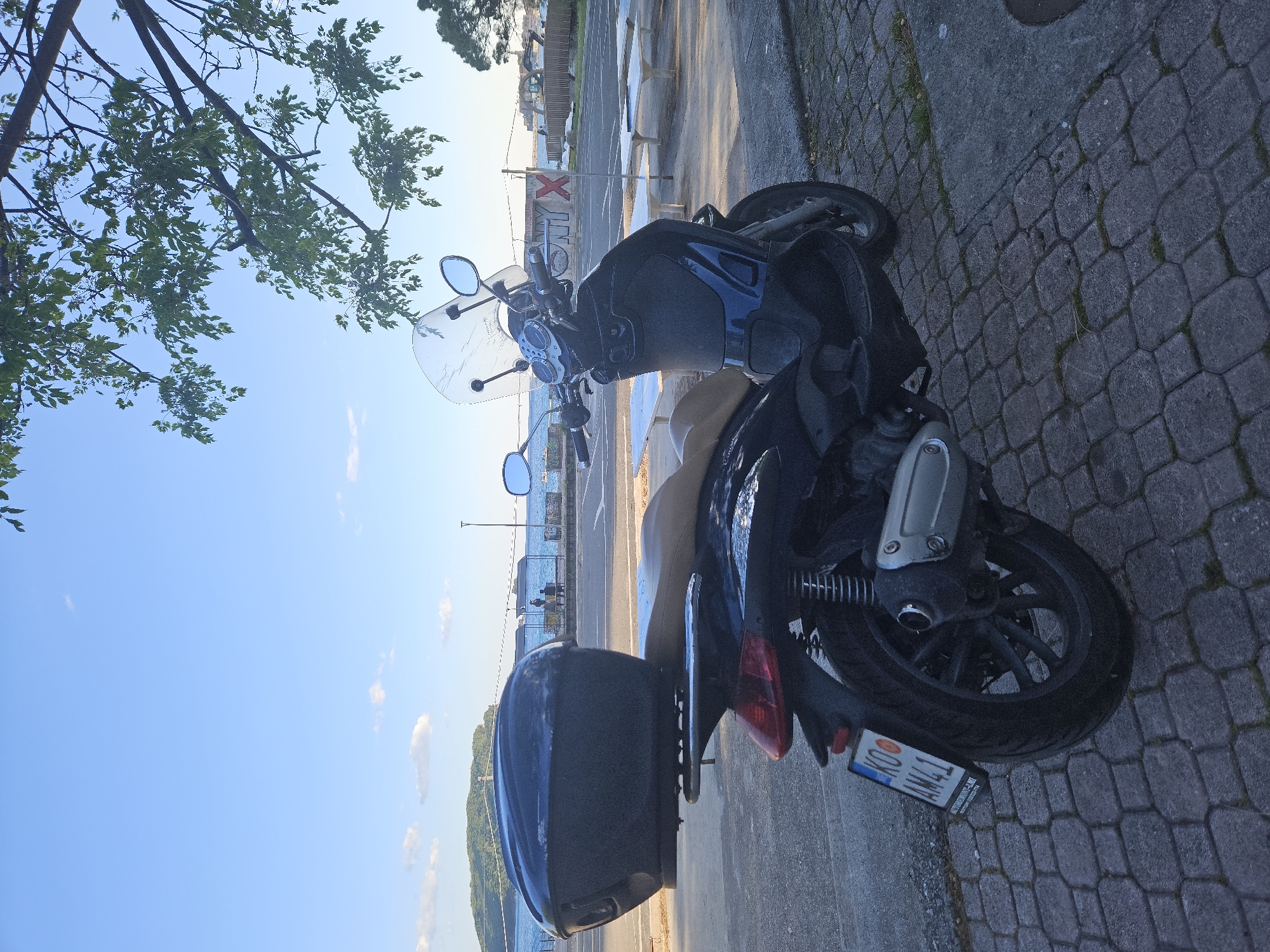 A vertical photograph, correctly oriented, captures a parked scooter near a body of water under a clear blue sky, likely in Budva, Montenegro.
In the foreground and left, lush green tree branches and leaves extend into the frame against the bright sky. The main subject is a dark-colored scooter, possibly black or dark blue, parked on a textured block-paved surface. It features a clear front windshield, rearview mirrors, and a black top case mounted on the rear rack. The scooter's license plate is visible, displaying "KO AMZ41" with "AUTOMOTO CLUB" written below.
To the right of the scooter, the paved surface transitions to a darker concrete path. In the mid-ground, behind the scooter, a road or larger paved area runs parallel to the water. Several light-colored concrete benches are intermittently placed along this path. A wall or barrier behind these benches features red graffiti or a sign with the letters "ONYX" and a large "X".
Beyond the road and benches, a vast body of water, likely the Adriatic Sea, stretches towards the horizon. On the distant horizon, rolling hills or mountains are visible under the expansive blue sky, which has only a few scattered white clouds. A few indistinct figures appear by the water's edge in the background, suggesting people enjoying the waterfront.
The lighting indicates a bright, sunny daytime, possibly morning or late afternoon, given the clear conditions and the way light hits the surfaces. The overall scene suggests a pleasant coastal setting.
