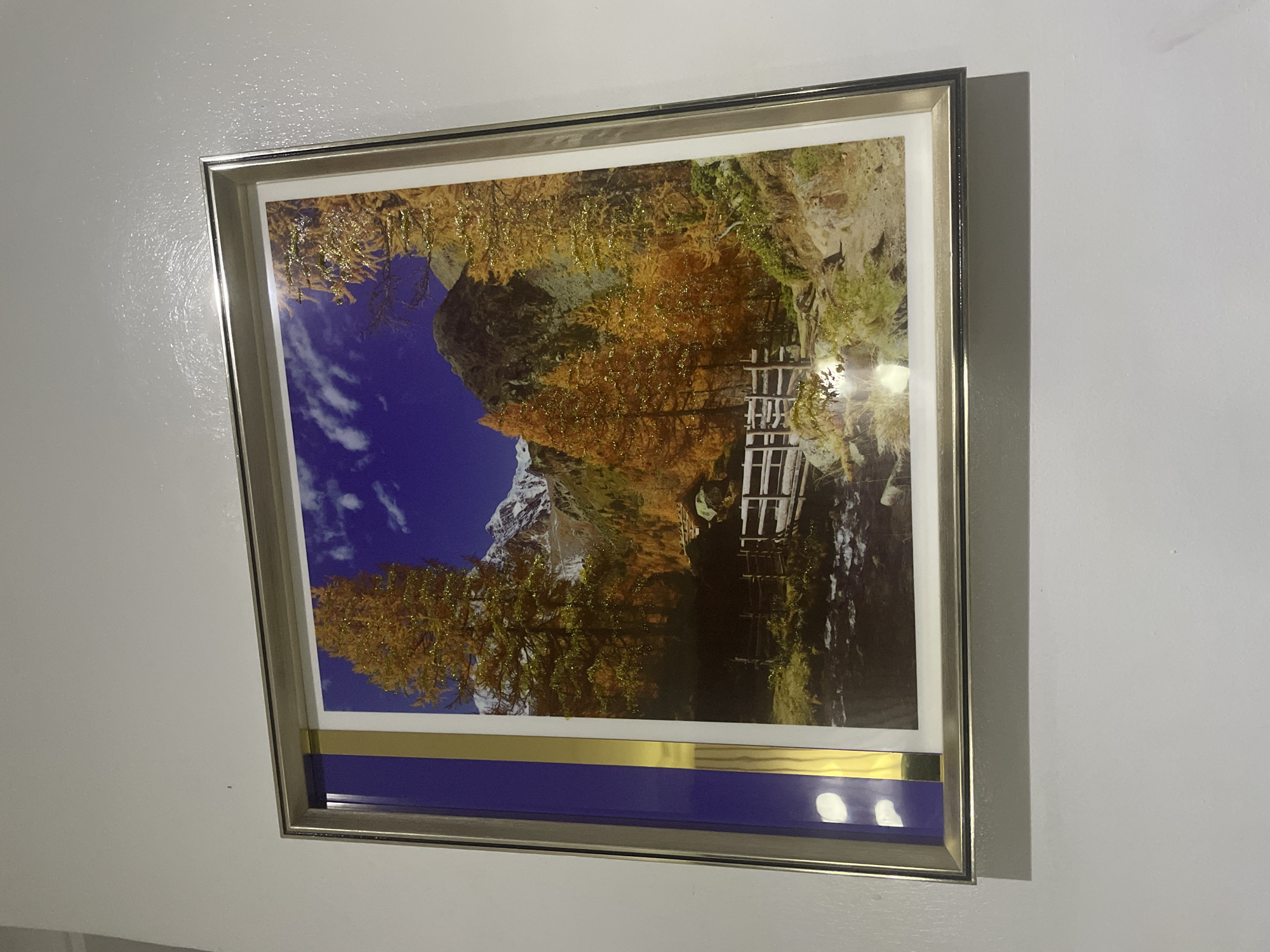 A framed print hangs on a white wall. The print depicts a mountain landscape in autumn with a blue sky. The print has a gold and blue border. The frame is silver. The location is unknown, and there are no people present.