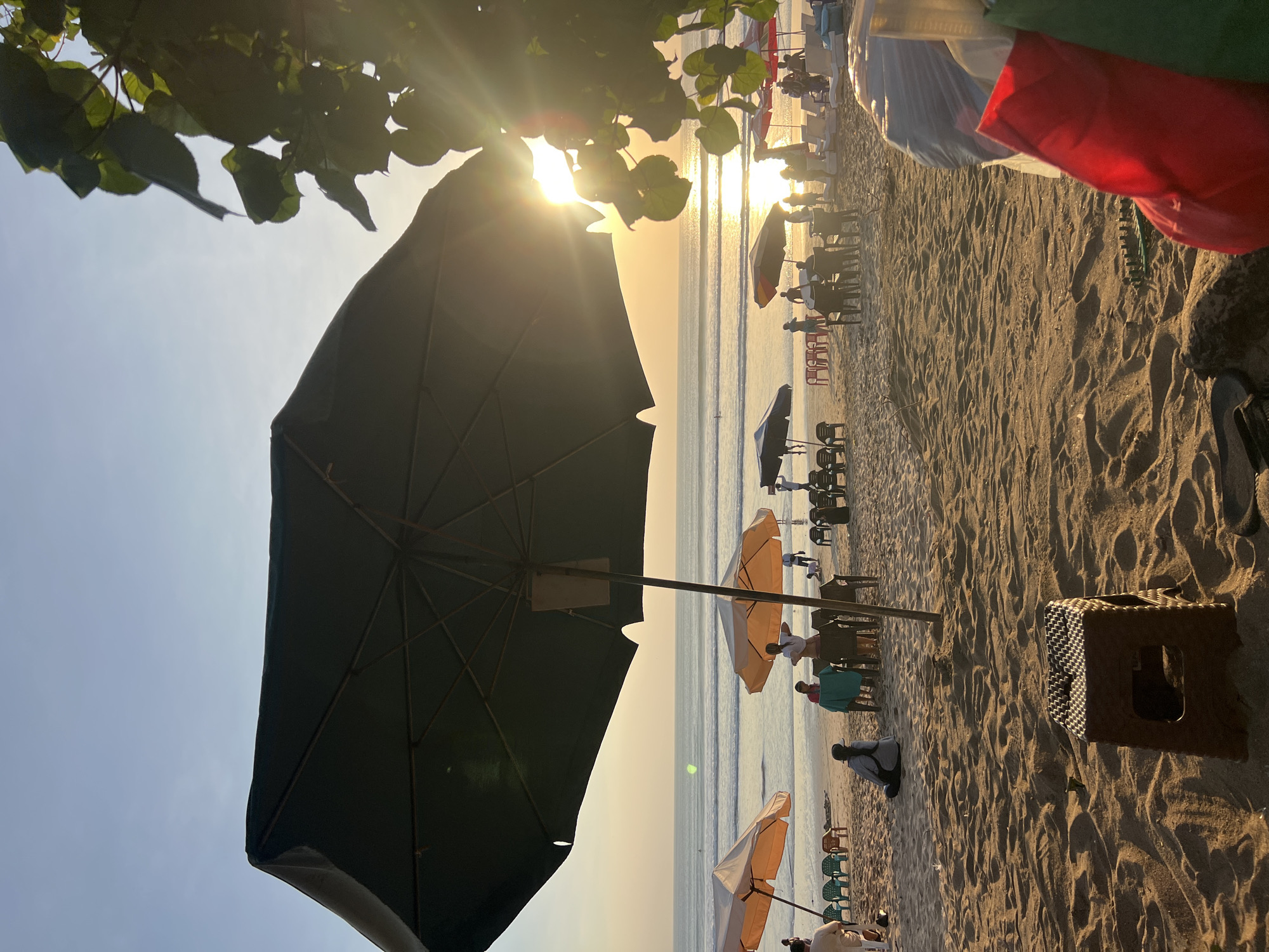 The image captures a serene sunset scene at Kuta Beach, Indonesia. The foreground is dominated by sandy terrain with a small, brown plastic stool in the center. In the midground, several beach umbrellas are set up, some with tables and chairs arranged underneath, indicating a relaxed seating area. People are visible scattered across the beach, some sitting, some standing, and some appearing to stroll along the shore.
The ocean stretches out to the horizon, with gentle waves rolling in. The setting sun casts a warm, golden glow across the water and the sky, creating a beautiful, tranquil atmosphere. In the upper right portion of the image, lush green foliage from a tree hangs into the frame, adding a natural element to the scene. The overall impression is one of a peaceful evening by the sea, with the soft light of the sunset enhancing the natural beauty of the location.
