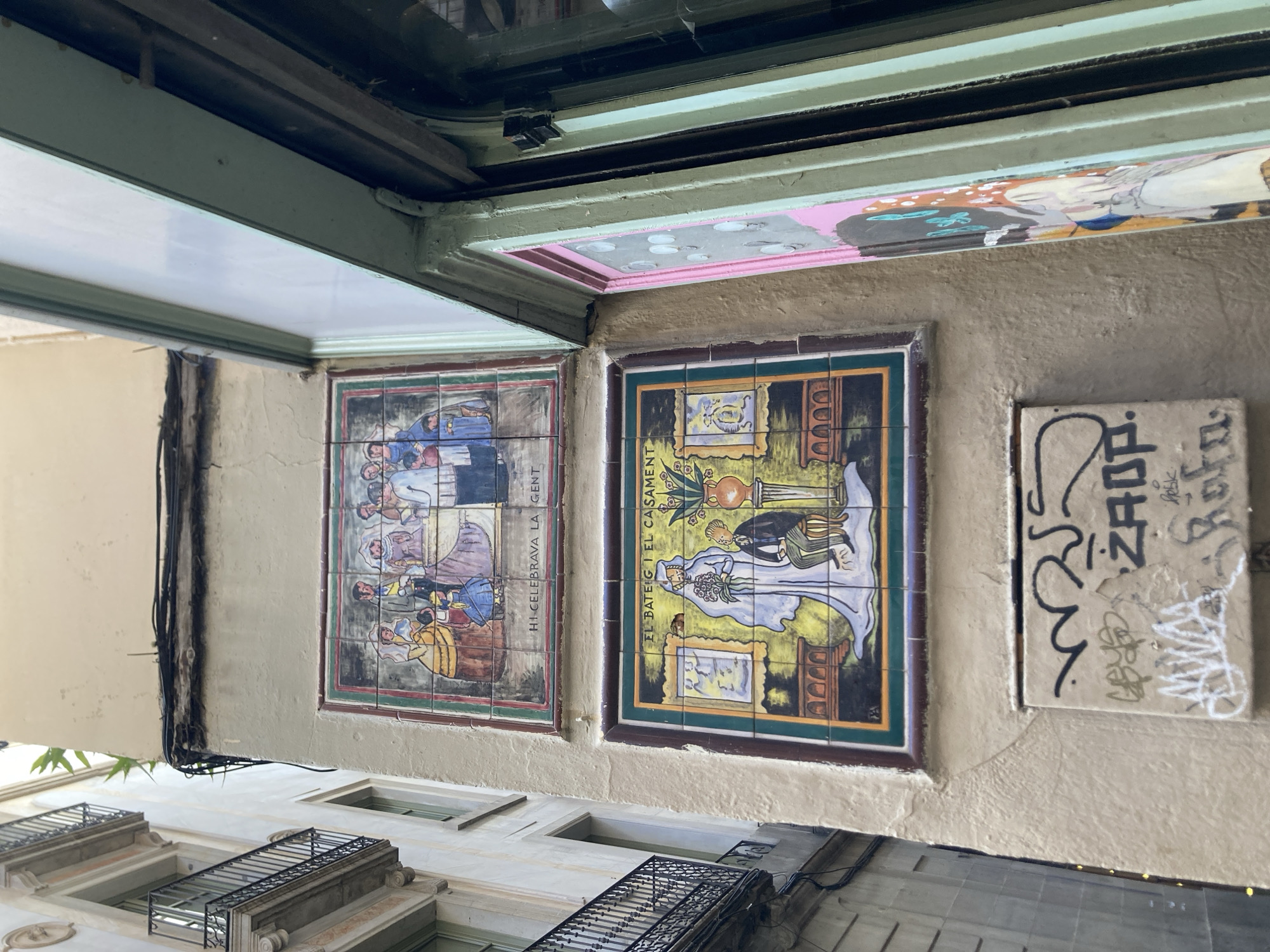 The image captures an outdoor scene in Barcelona, Spain, showcasing decorative ceramic tiles on a building's facade. The perspective is from below, looking upwards.
The primary subjects are two rectangular ceramic tile murals. The left mural depicts a group of people dressed in historical or traditional attire, possibly engaged in a communal gathering or ceremony. Several figures are visible, rendered in a colorful, illustrative style. Below this mural, partially obscured, is text that reads "HI. CELEBRAVA LA GENT".
The right mural features a scene that appears to depict a wedding. A bride in a white wedding dress holding a bouquet is central, alongside a groom in formal attire. Behind them, a decorative urn or vase is prominent. This mural also has text below it, reading "EL BATEIG I EL CASAMENT".
To the right of these murals is a smaller, rectangular tile with graffiti art. The graffiti is in black and white, with bold lines and stylized lettering, obscuring any potential original details on this tile.
The building architecture visible includes balconies with wrought-iron railings on lower levels, suggesting residential or commercial spaces. The overall setting appears to be an urban environment, possibly a narrow street or courtyard given the close proximity of buildings. The lighting suggests it is daytime, though the weather is not explicitly discernible. The condition of the murals and the presence of graffiti indicate an aged but still vibrant urban landscape.