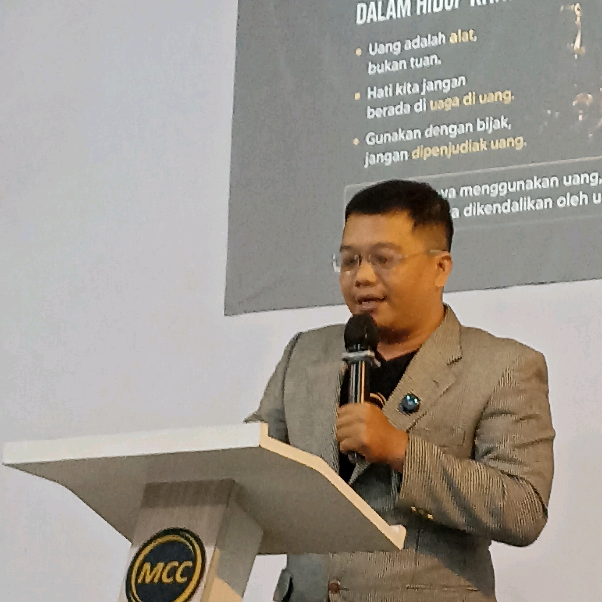 A man wearing glasses and a grey pinstripe suit stands behind a white podium speaking into a microphone. A logo with the letters "MCC" in a circle is visible on the podium. A projected image is visible behind the man, displaying text in Indonesian. The text includes bullet points that translate to: "Money is a tool, not a master." "Let's not be enslaved by money." "Use wisely, don't gamble with money." "When using money, let it be controlled by you." The background appears to be a plain wall. The lighting suggests an indoor setting, possibly a conference room or auditorium. The man's posture and facial expression indicate he is actively engaged in a presentation. The overall scene suggests a formal event or educational seminar. There is no discernible connection to the city of Magelang, Indonesia, within the image itself.