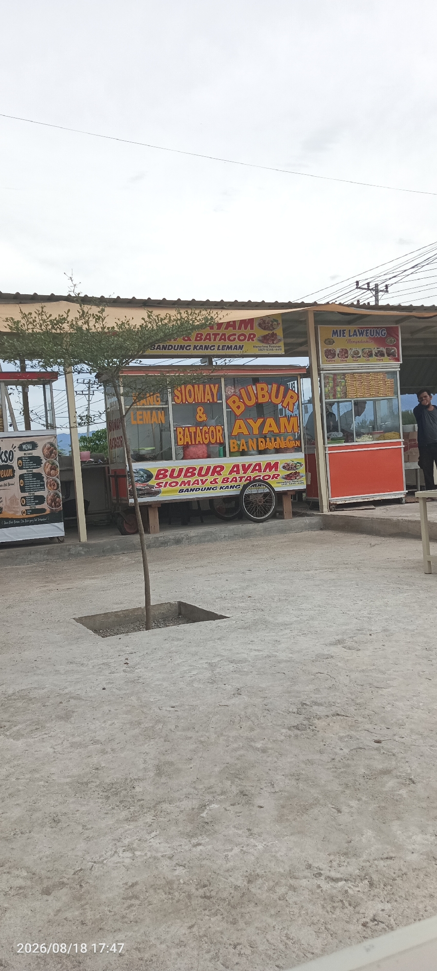 An outdoor food stall area is depicted, with a concrete ground and a light-colored overcast sky. Several food carts are visible, each with colorful signage advertising various dishes. One cart prominently displays "BUBUR AYAM SIOMAY & BATAGOR BANDUNG KANG LEMAN," with images of food items. Another cart has a sign for "MIE LAWEUNG." To the left, a cart labeled "AKSO KEUN" is partially visible with a menu board. A small tree grows from a square opening in the concrete in the foreground. In the background, a person in a dark shirt and pants stands near a red food stall. Electrical wires are strung overhead, supported by poles. A table is visible on the far right. The timestamp "2026/08/18 17:47" is present in the lower left corner of the image. The scene appears to be a commercial area, likely a roadside food court in Cotcut, Indonesia.