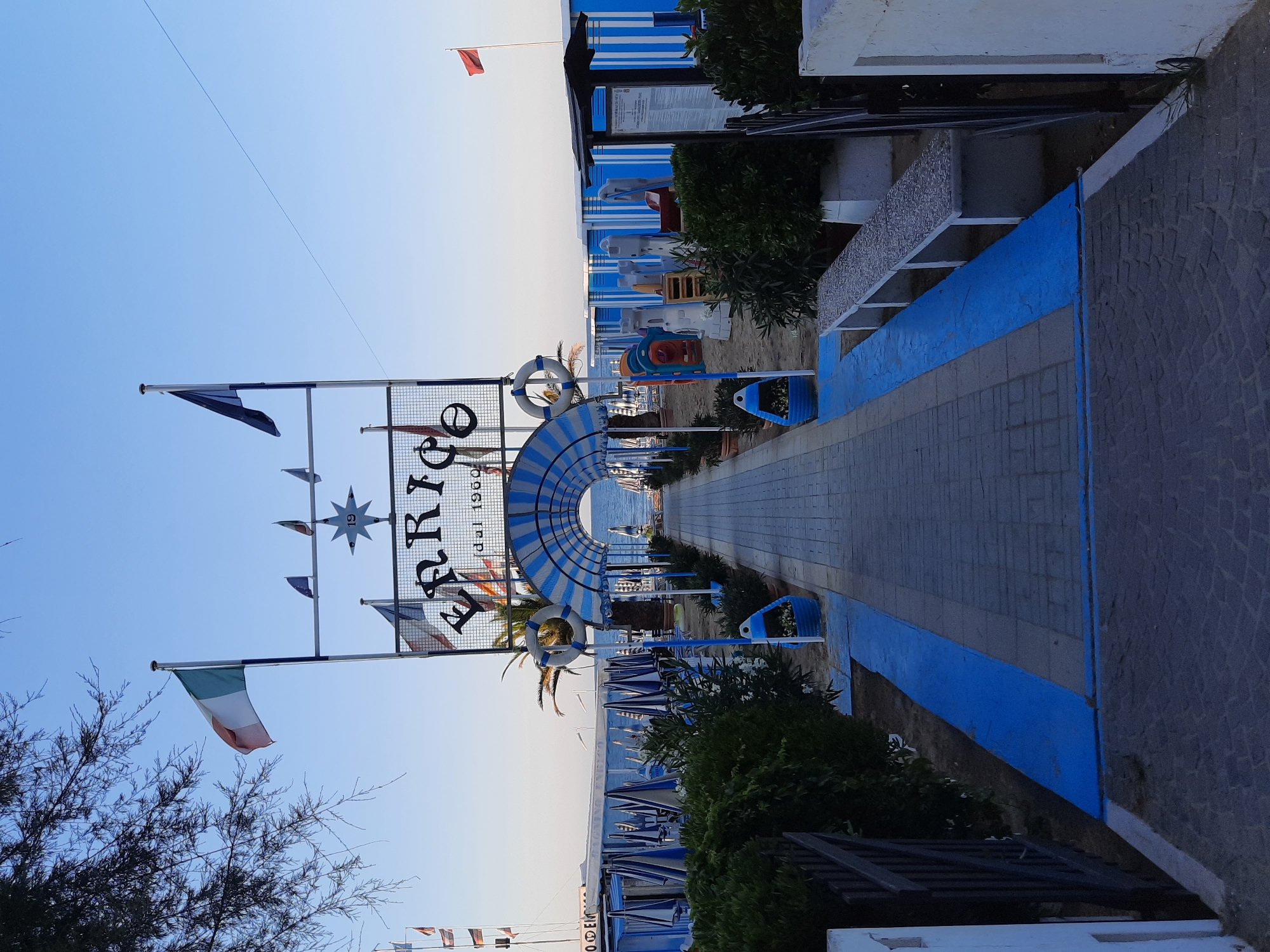 The image shows an outdoor scene in Pesaro, Italy, likely a beach resort. A paved walkway, bordered by blue painted areas, leads towards the sea. The walkway is covered by a blue and white striped archway. On either side of the walkway are rows of beach huts, predominantly blue and white, and green bushes. A sign at the entrance to the archway reads "ERRICO" and "dal 1960". Several flags are displayed on poles, including the Italian flag. There are also children's play structures visible on the sand to the right. The sky is clear and blue, indicating daylight.