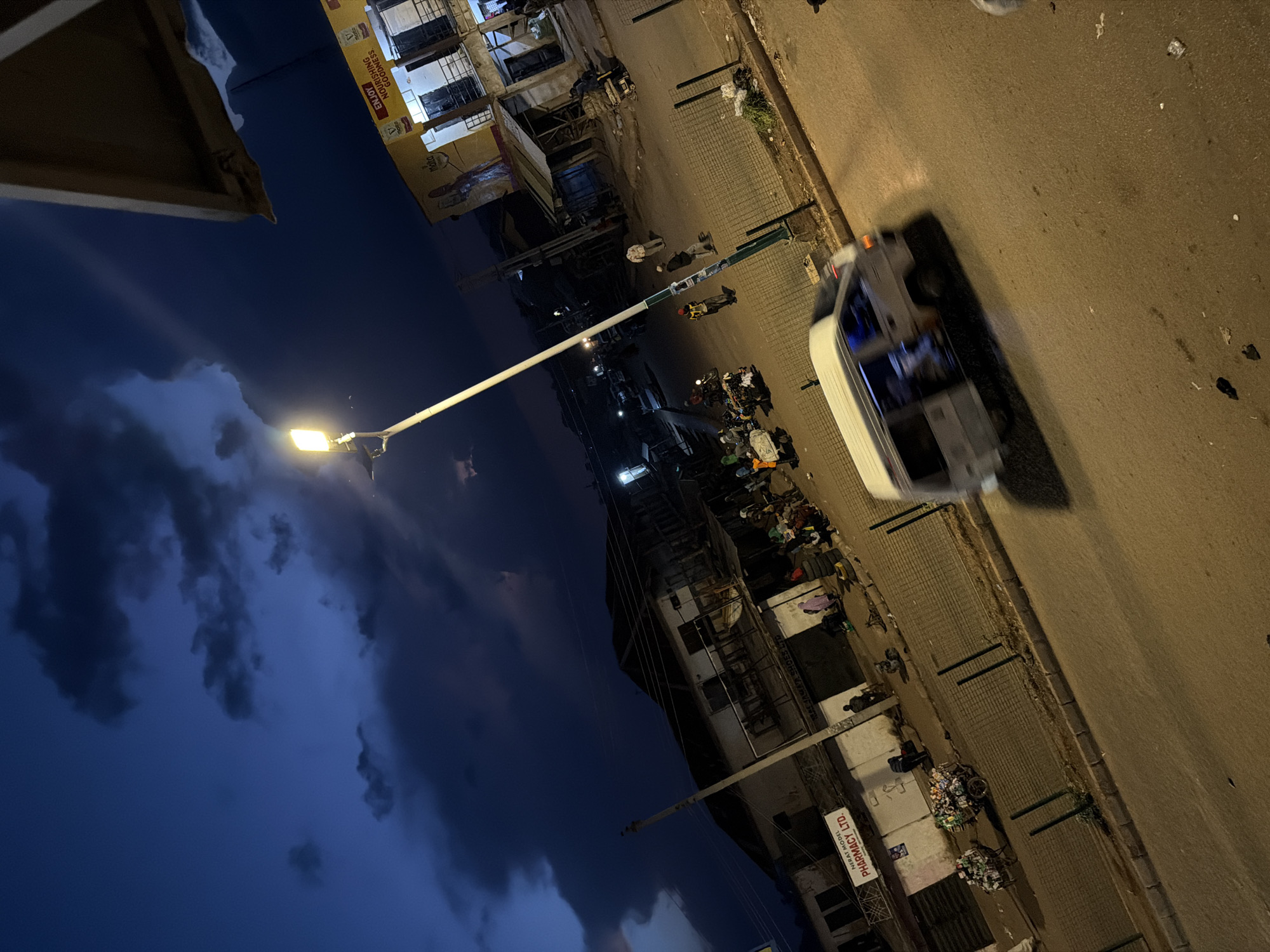 The image depicts a street scene at dusk in Bukuru, Nigeria. A white van is captured in motion, its doors open, with people visible inside. Several individuals are standing on the sidewalk and street, and two carts loaded with goods are present. Buildings line the street, one of which has a sign for "NERAT MODEL PHARMACY LTD." A street lamp illuminates the area, and the sky is a deep blue with dark clouds. The overall atmosphere suggests a busy market or commercial district at the end of the day.