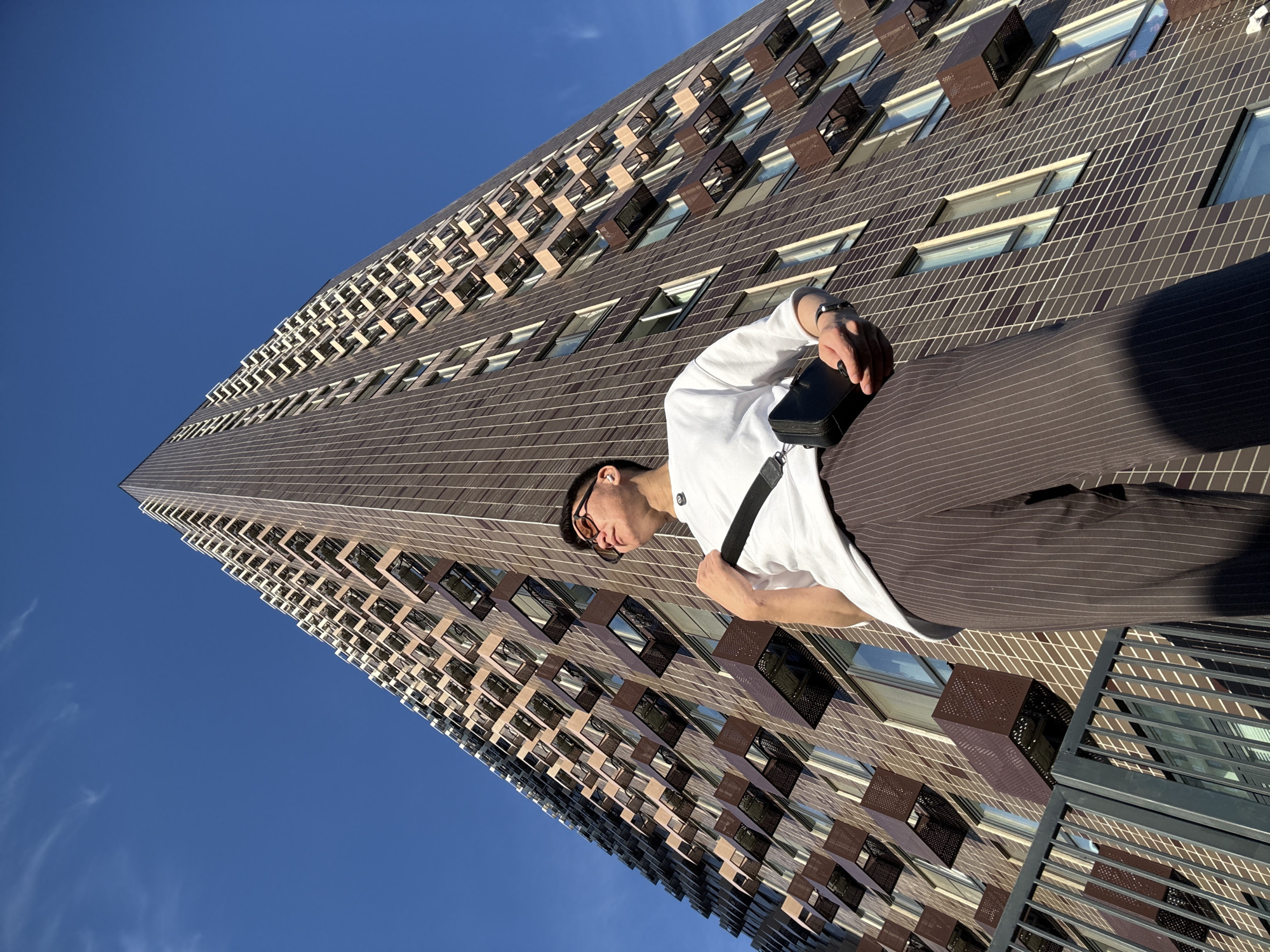 An individual, appearing to be male, stands outdoors against a backdrop of a tall, modern apartment building with a distinctive patterned facade and numerous balconies. The building is a dominant element, reaching towards a clear blue sky. The individual is looking upwards and slightly to the right, with sunglasses on and a small black bag slung across their chest. They are wearing a white t-shirt and dark, pinstriped trousers. The perspective of the shot is from below, emphasizing the height of the building and the individual's positioning in relation to it. The scene appears to be a public space, possibly a residential area or courtyard, given the presence of a railing in the foreground. The lighting suggests it is daytime, with the sun casting some shadows. Based on the architectural style and design of the building, it is characteristic of contemporary residential complexes found in Moscow, Russia.