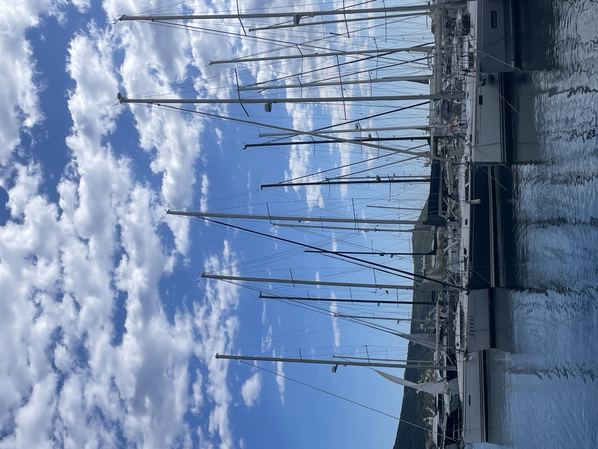 The image shows a harbor scene in Meljine, Montenegro. Multiple sailboats are moored at a dock, their masts reaching towards a cloudy blue sky. The water reflects the hulls of the boats and the sky above. A green hillside is visible in the background.