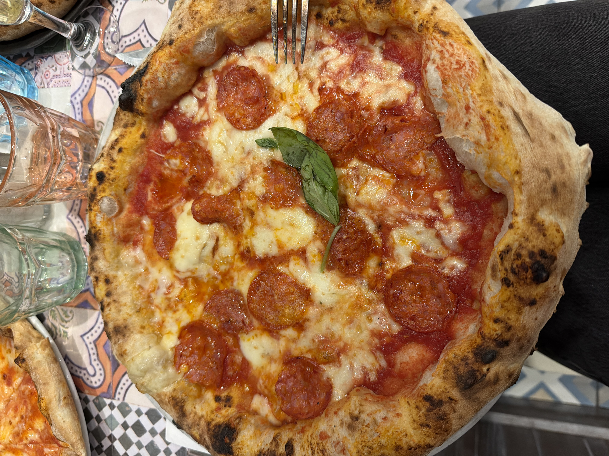A close-up, overhead shot shows a pepperoni pizza on a white plate. The pizza has a thick, puffy crust with some charred spots, and is topped with tomato sauce, melted mozzarella cheese, and round slices of pepperoni. A sprig of fresh basil rests on top of the pizza. To the right of the pizza, a silver fork with three tines is visible. In the background, out of focus, are two glasses of drinks, one clear and one pink, and a wine glass. The pizza is placed on a table with a colorful tiled pattern. The scene appears to be in a restaurant in Milan, Italy.