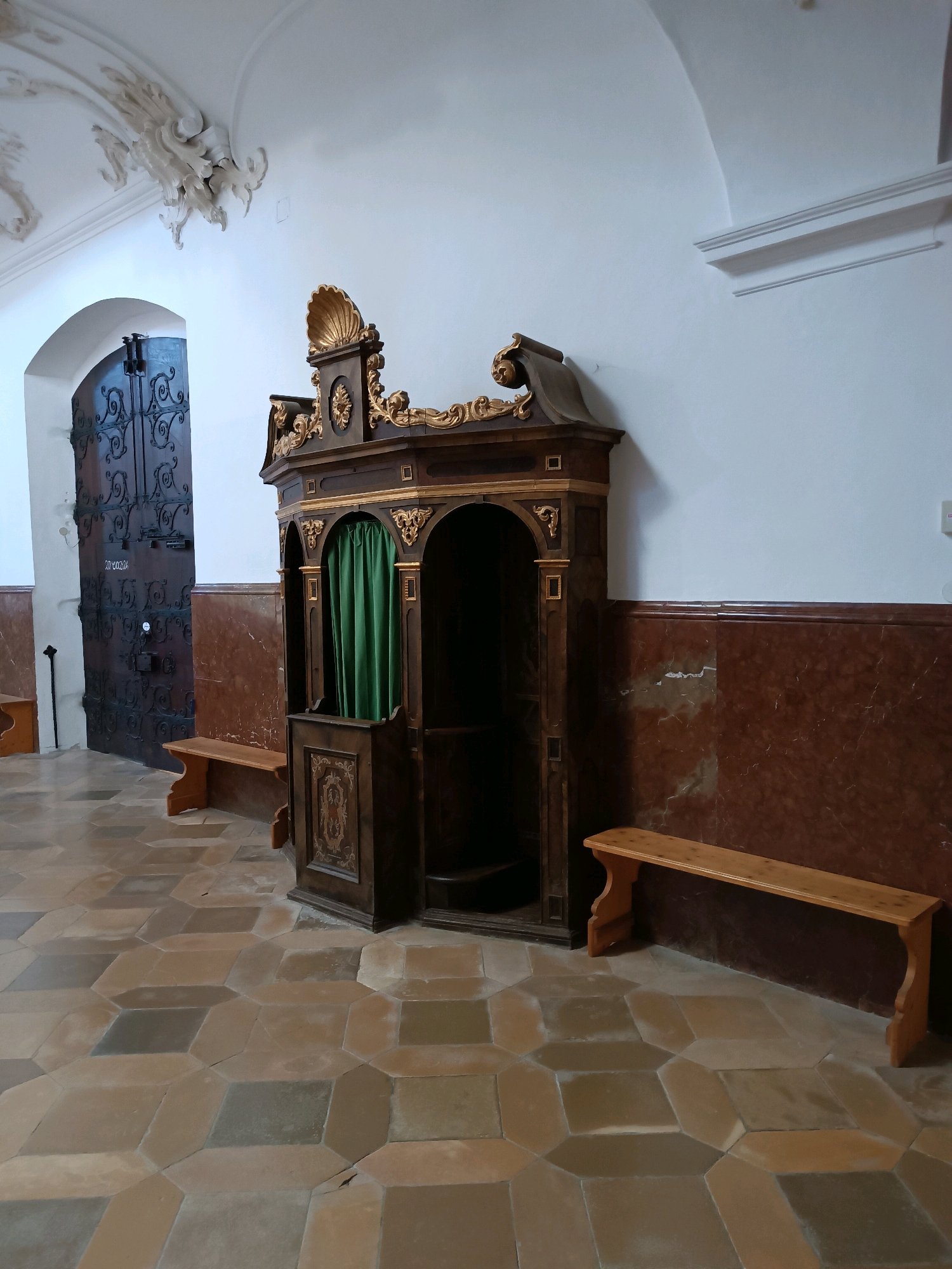 The image depicts the interior of what appears to be a church or religious building. In the center, a dark wooden confessional booth with ornate gold detailing and a green curtain is the main subject. To the left of the confessional is a large, dark metal double door with intricate scrollwork. Two simple wooden benches are positioned in front of the confessional and to the left of the door. The floor is tiled with a geometric pattern of light brown and greyish-brown hexagonal and diamond shapes. The walls are white, with a wainscoting of reddish-brown marble or patterned material. Ornate white stucco decorations are visible on the ceiling and upper wall on the left side of the image. The lighting appears to be natural, coming from an unseen source, casting soft shadows. No people are present in the scene.