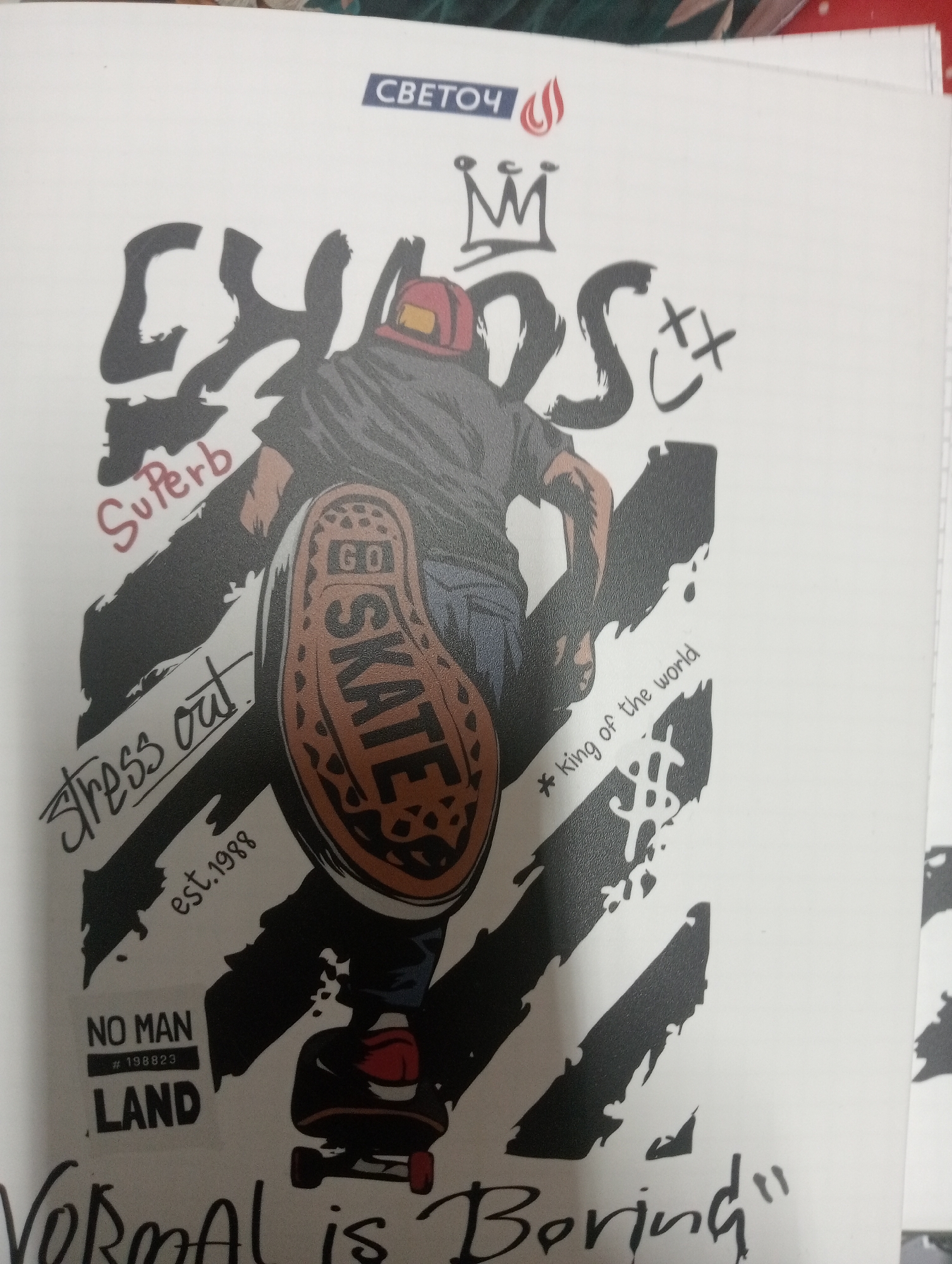 This is a photograph of a notebook page featuring a stylized graphic illustration. The illustration depicts a person in mid-action on a skateboard, viewed from behind and slightly from the side. The person is wearing a red helmet and dark clothing. The sole of the skateboarder's shoe is prominently displayed and features the word "SKATE" in bold lettering, with "GO" above it.
Other text elements in the illustration include:
- "CHAOS" in large, distressed lettering across the top.
- "SuPerb" written in a smaller, cursive font below "CHAOS."
- "stress out." in a handwritten style.
- "est. 1988" positioned below "stress out."
- "* King of the world" to the right of the skateboarder.
- "NO MAN" and "LAND" with a small hashtagged number "# 198823" in a rectangular block.
- A crown graphic above "CHAOS."
- The word "СВЕТОЧ" in a blue rectangle at the top, with a flame symbol next to it.
- The phrase "vormal is Boring" is written in large, handwritten script at the bottom of the page.
The background of the illustration consists of bold, diagonal black stripes on a white surface. The photograph is taken from an overhead perspective, with the notebook page angled. The lighting appears to be natural, possibly from a window, casting some subtle shadows. The image shows a portion of the notebook's binding on the right side.
There is no visible information or context within the image that would indicate its location in the city of Aktobe, Kazakhstan.