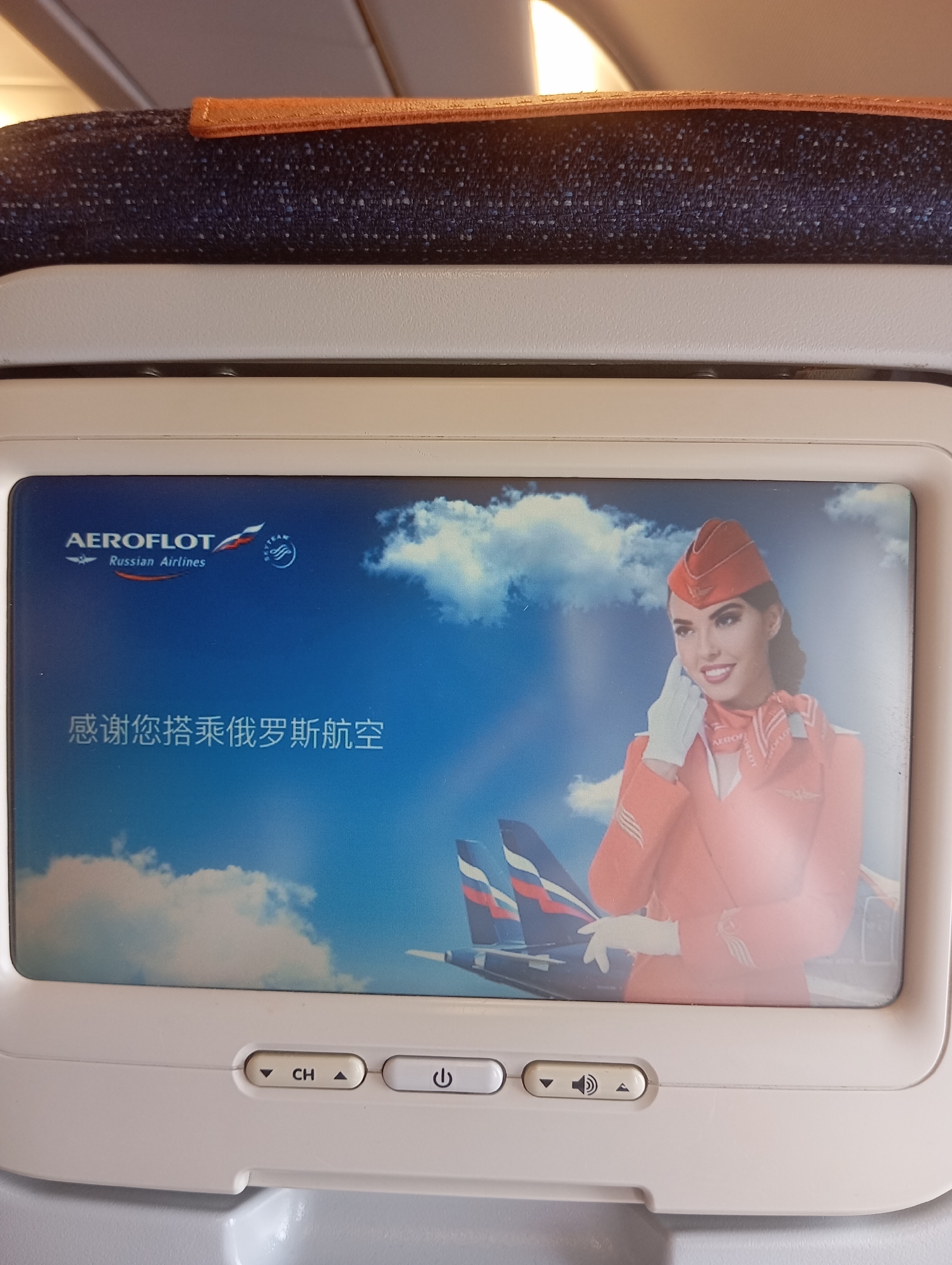 An Aeroflot Russian Airlines screen displays a flight attendant in an orange uniform with a red hat, smiling and holding a white glove to her cheek. The background features a bright blue sky with scattered white clouds and two aircraft tails. Below the image, a panel shows control buttons for channel selection, power, and volume. The screen is positioned within an airplane cabin. The context of Tambon Mai Khao, Thailand, is not discernible from the image itself.