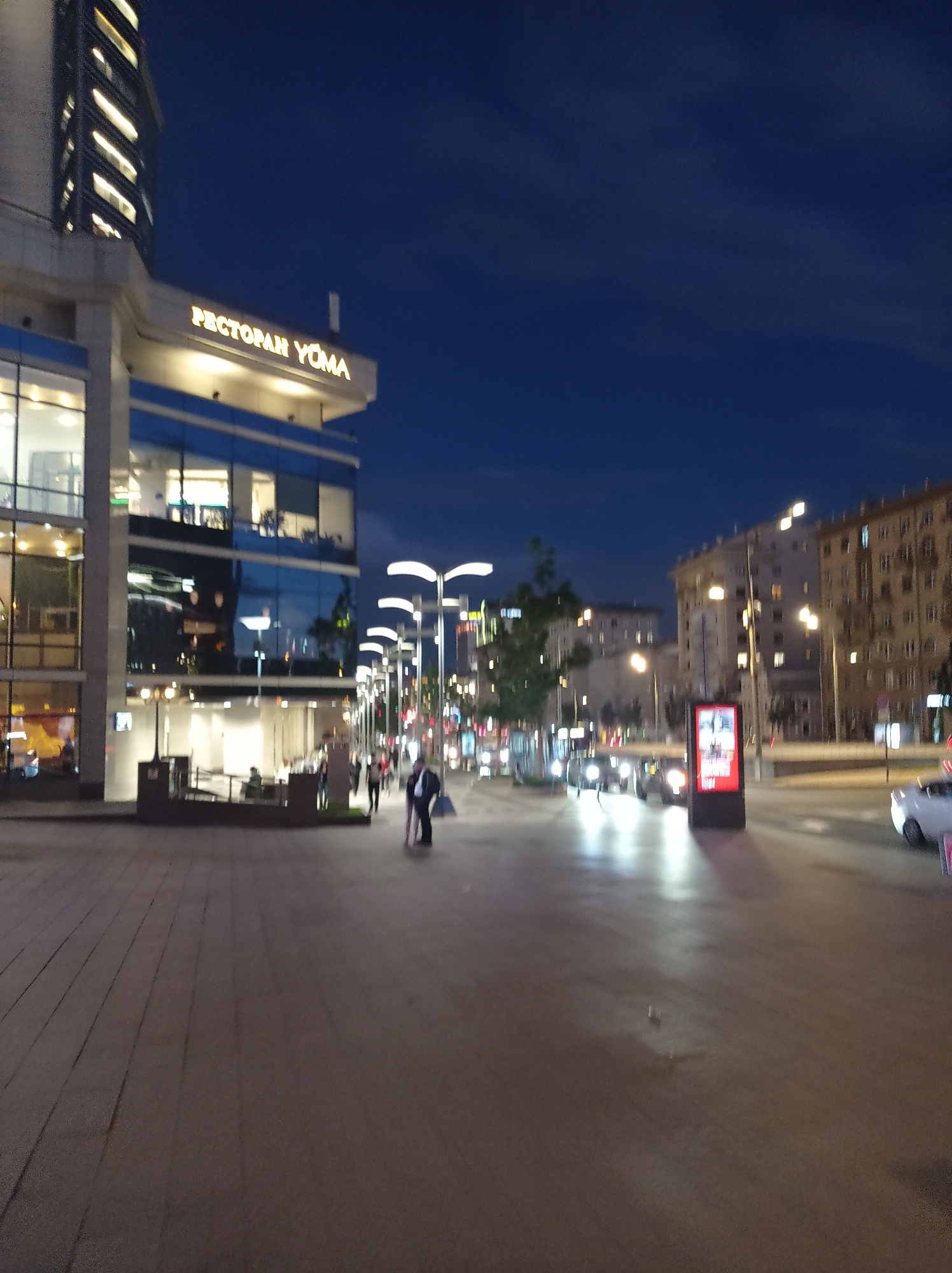 The image depicts a night scene on a city street in Moscow, Russia, likely near a commercial building. In the foreground, a paved walkway or plaza is visible, with a few people walking. To the left, a modern building with large glass windows and illuminated interiors stands prominently. A sign above the entrance reads "РЕСТОРАН YOMA" (RESTORAN YOMA) in illuminated letters. Further down the street, a row of streetlights with distinctive curved designs illuminates the area. Vehicles are present on the road, and several residential or office buildings with illuminated windows line the opposite side of the street. A digital billboard displaying an advertisement is also visible on the right side of the road. The sky is a deep blue, indicating it is dusk or early night. The overall impression is of a well-lit urban environment in the evening.