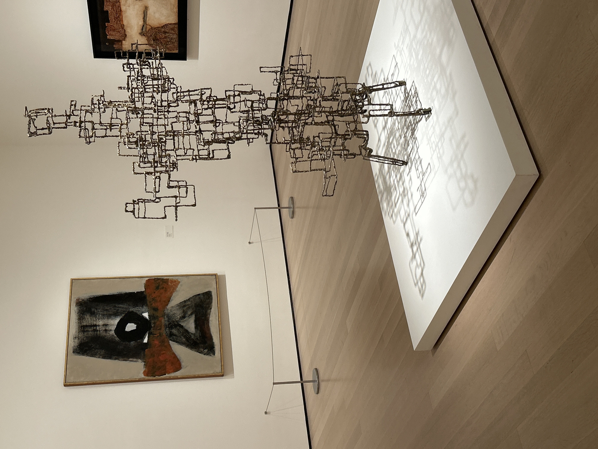 The image displays an interior view of an art gallery or museum, likely in New York, USA, showcasing several abstract artworks. The central and most prominent subject is a large, metallic abstract sculpture composed of numerous interconnected square and rectangular forms. This intricate, three-dimensional structure appears to extend horizontally across the space, casting a detailed and complex shadow onto a white, rectangular display plinth positioned on the light-toned hardwood floor. The shadow precisely mirrors the angular and fragmented nature of the sculpture.
Two abstract paintings are mounted on the white wall. Above the sculpture, a painting with earthy tones and textures is framed in black, partially visible. Below the sculpture, a larger abstract painting, set in a light wooden frame, features bold black and white brushstrokes with a distinctive orange, hourglass-shaped element at its center. Thin wires with small metallic anchors are visible on the floor, possibly delineating a viewing boundary or supporting elements of the display.
The setting is a well-lit indoor space, indicative of an exhibition hall, with artificial lighting accentuating the artworks. The absence of people emphasizes the static display of the art. No discernible text is legible on the artworks or any accompanying labels.