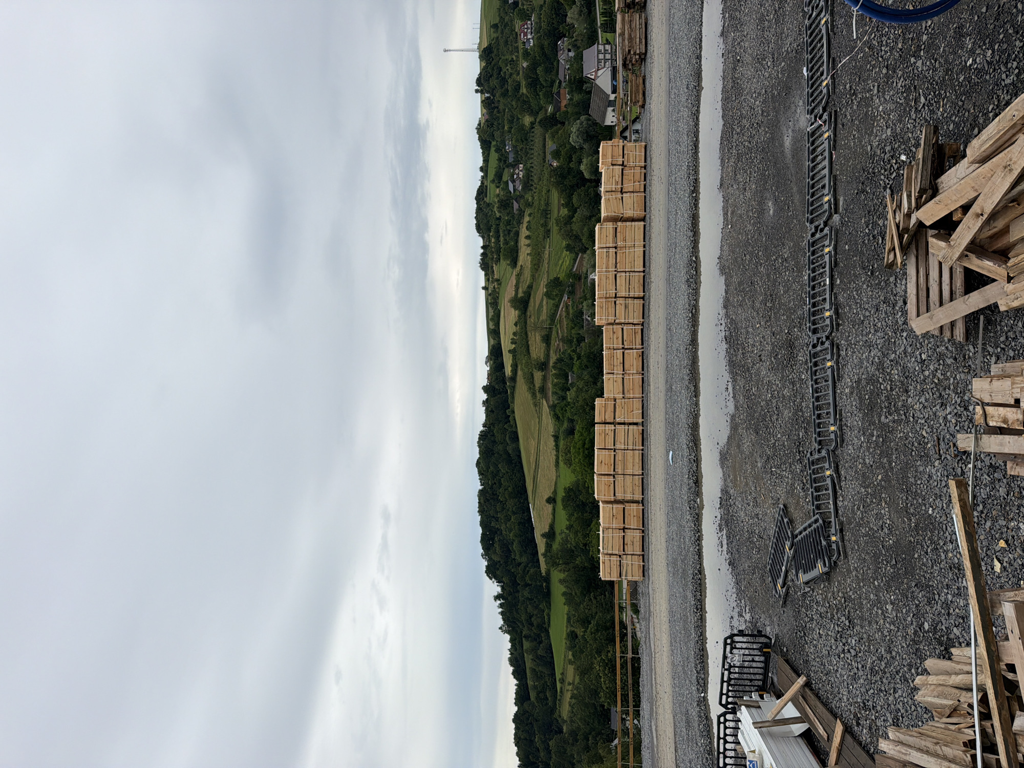 The image shows an outdoor scene with a hilly, green landscape in the background, dotted with houses and trees. In the foreground, there is a gravel area with several stacked wooden crates. To the right of the crates, there are also piles of discarded wooden planks and some black plastic barriers. A water puddle is visible on the gravel. The sky is overcast, suggesting it is daytime, possibly in the morning or late afternoon. The overall impression is that of a storage or construction area adjacent to a rural landscape. There is no visible text in the image.