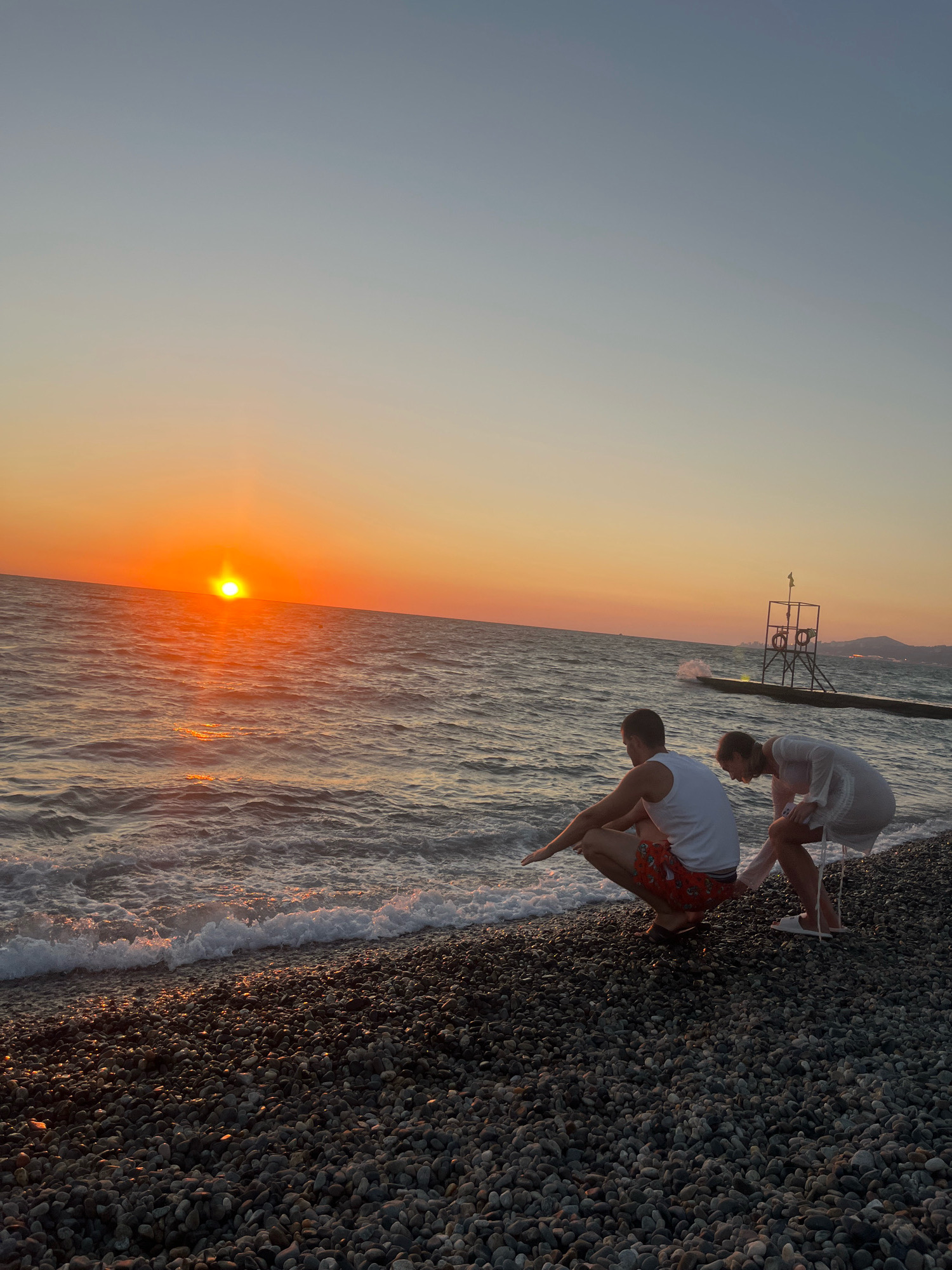 A man and a woman are crouched on a pebble beach near the water's edge, facing the sea at sunset. The man, wearing a white tank top and patterned shorts, extends his left arm as if touching the water. The woman, in a white dress, is also leaning forward, her hands near the shore. The sun is setting over the ocean, casting a warm orange glow across the sky and reflecting on the water. In the background, a pier extends into the sea with a lifeguard tower on it. The scene suggests a peaceful evening on the coast of Sochi, Russia.