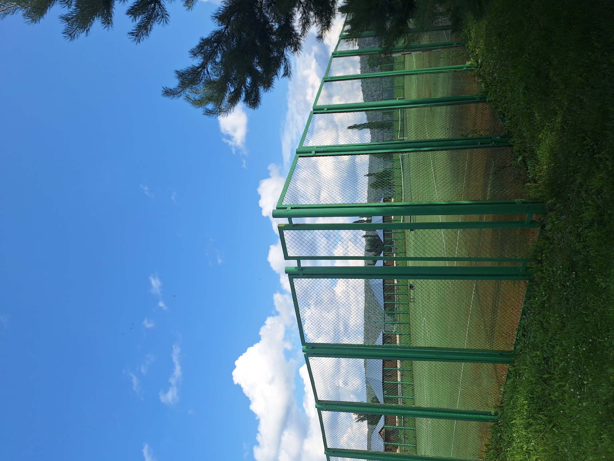 A bright, sunny day with a clear blue sky filled with scattered white clouds. In the foreground, a green metal chain-link fence surrounds a tennis court. The court's surface appears to be a mix of green and reddish-brown material. Beyond the fence, several small wooden buildings with dark roofs are visible, set against a backdrop of rolling green hills. A few pine tree branches frame the top right corner of the image. The overall scene suggests an outdoor recreational area, possibly a sports complex or a resort, located in or near the city of Atamanovka, Russia. There are no people visible in the image.