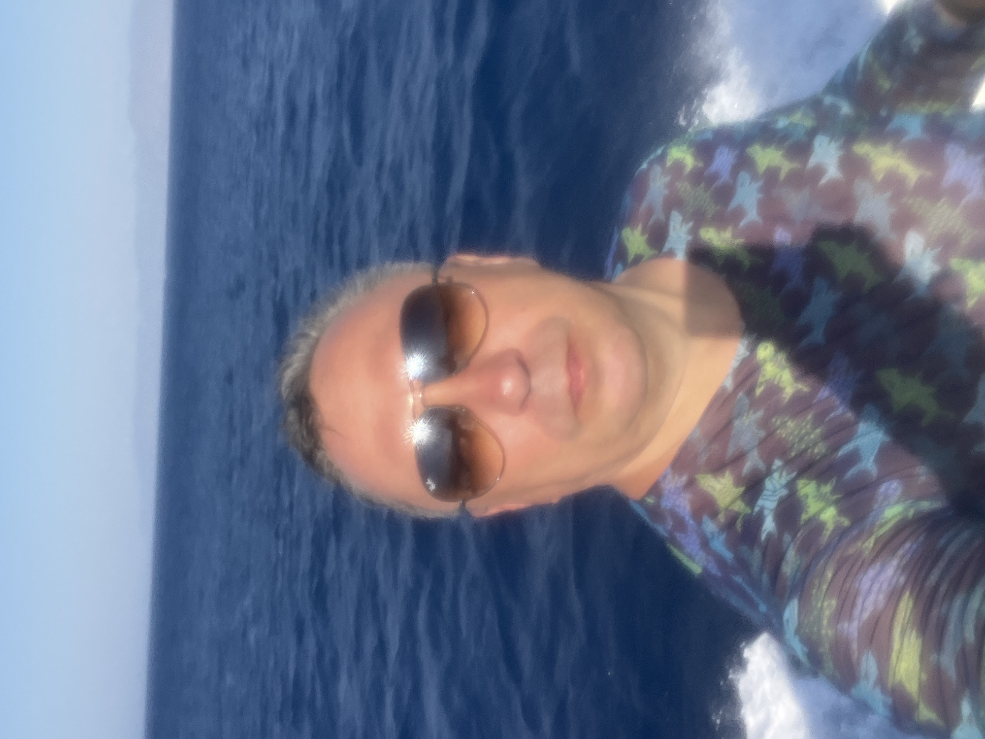 A man is taking a selfie while on a boat at sea. He is wearing sunglasses and a t-shirt with a pattern of sharks. The water is blue and there is a wake behind the boat. The sky is clear and blue. The location is the sea near Neo Chorio, Cyprus.