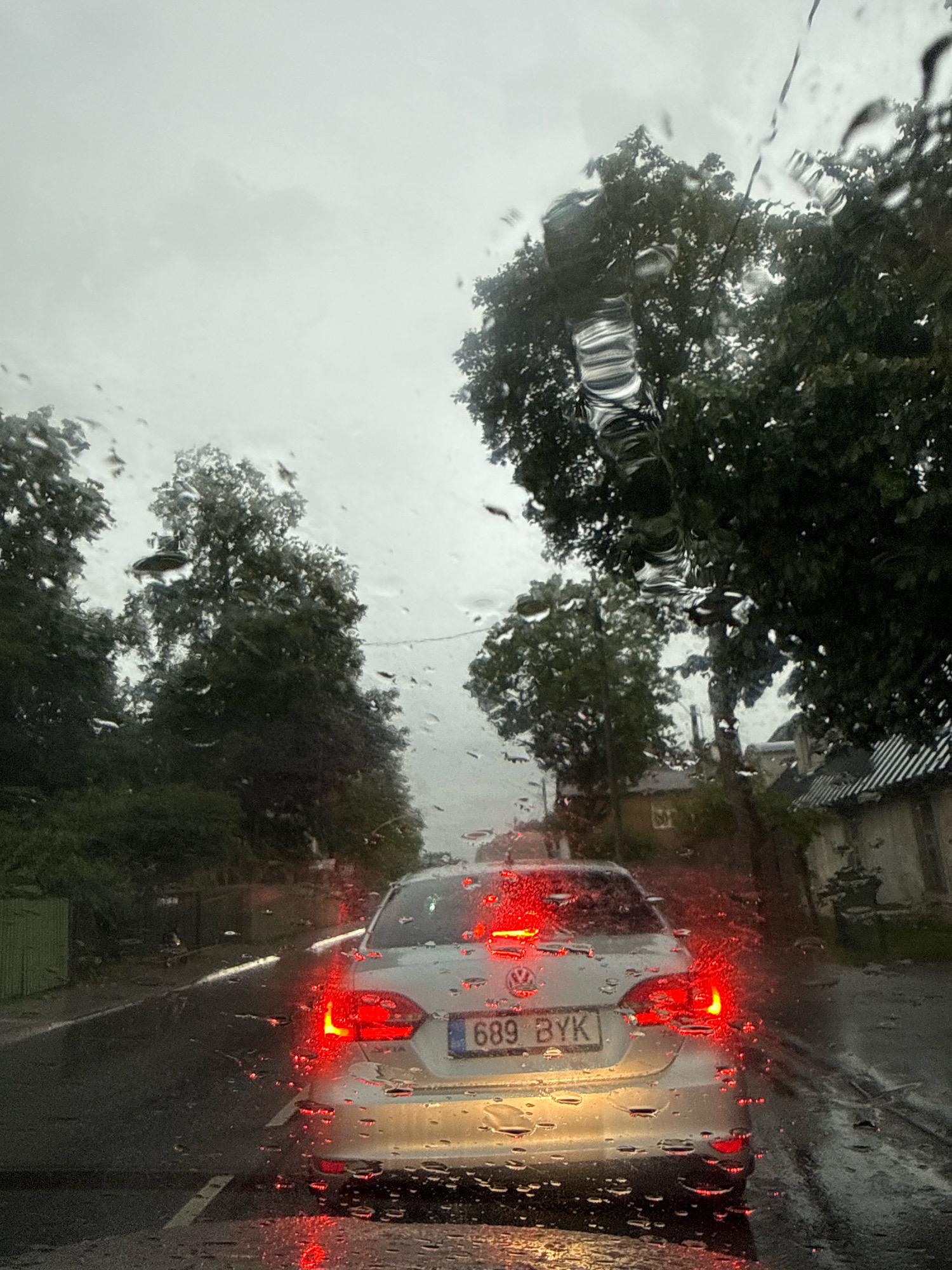 The image, taken from inside a vehicle, depicts a street scene in Tallinn, Estonia, on a rainy day. Water droplets are visible on the windshield, distorting the view of the surroundings. The focus is on the rear of a silver Volkswagen sedan with the license plate "689 BYK" driving on a wet road. The car's brake lights are illuminated, casting a red glow on the road and the surrounding environment. Tall trees with dark green foliage line the street. In the background, residential buildings are partially visible. The sky is overcast and grey, contributing to the somber mood of the scene. The overall impression is that of a typical overcast day in Tallinn, with traffic moving through residential streets.